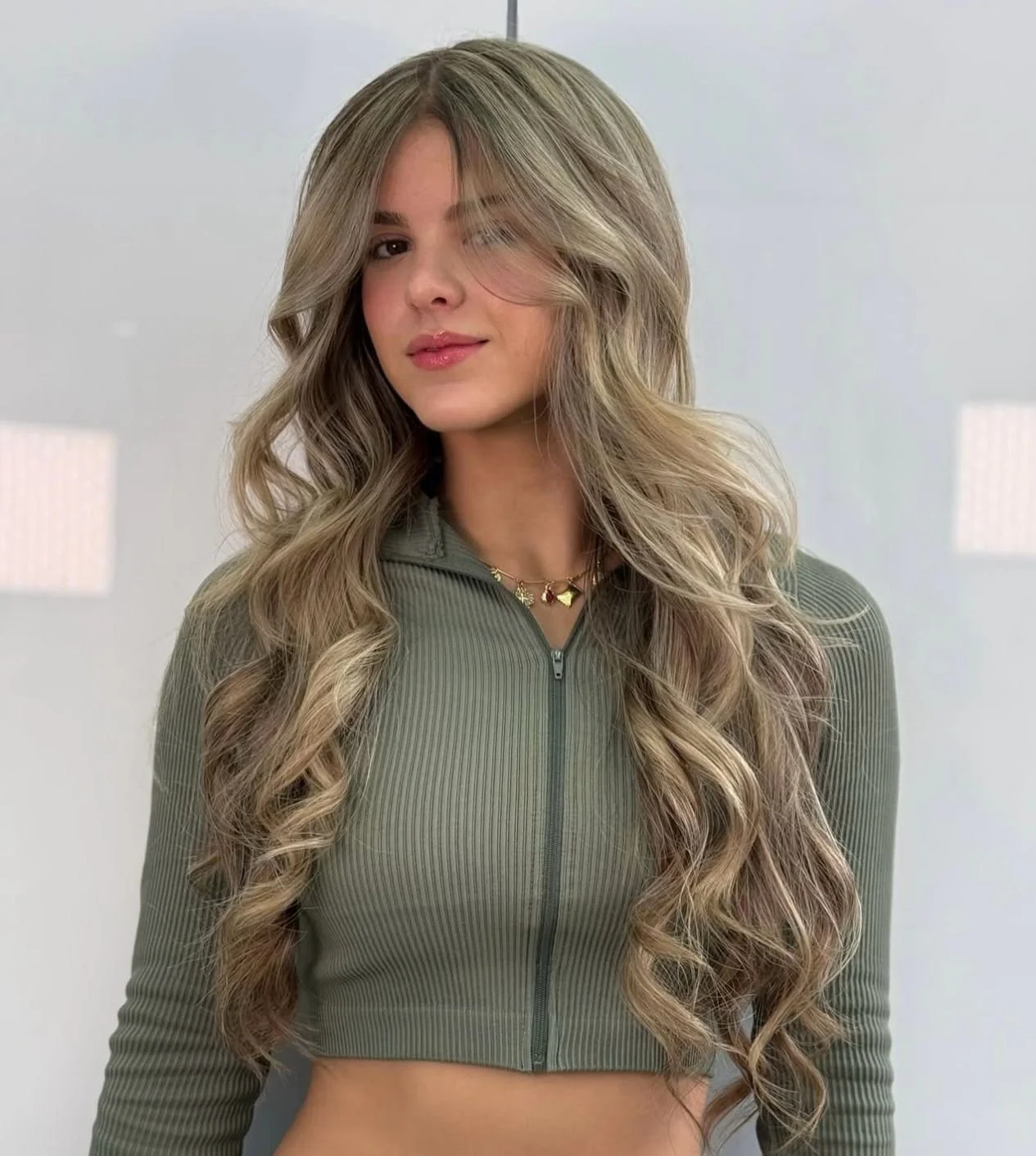 A young woman with long, wavy blonde hair wearing a green zip-up top and a gold necklace, looking at the camera with a slight smile and winking.