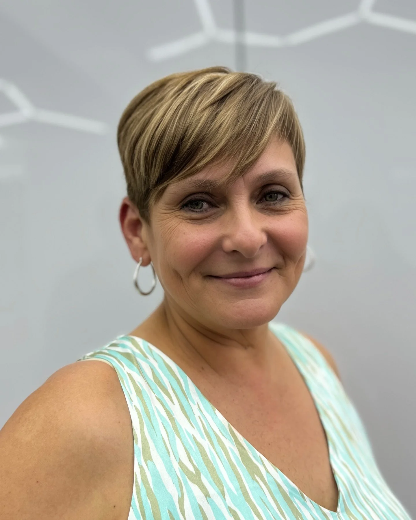 A woman with short light brown hair, wearing a sleeveless light green and white patterned top, smiling slightly against a plain light gray background.