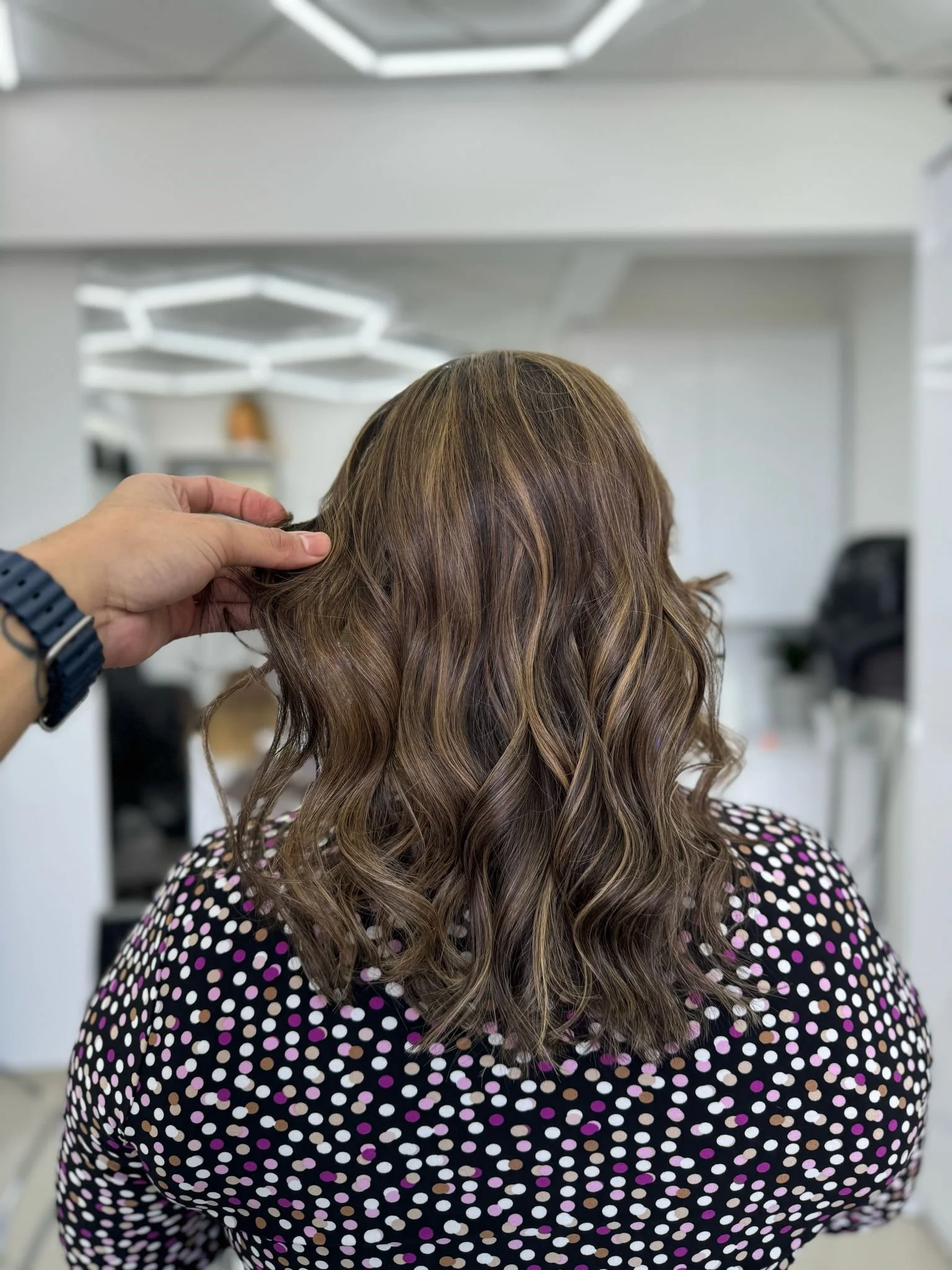 Back view of a woman with curly brown hair with blonde highlights, wearing a black dress with multicolored polka dots, in a salon or studio.