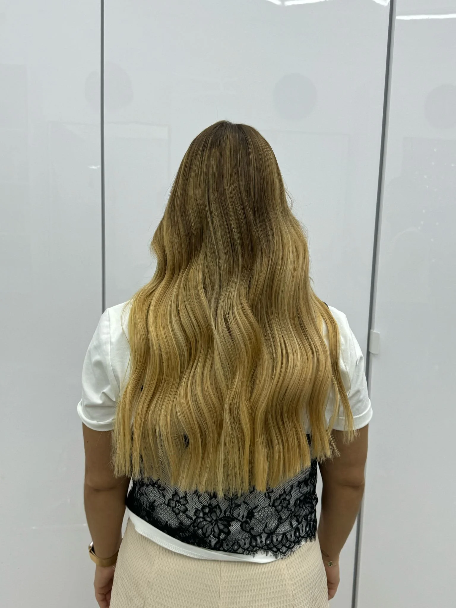 Back view of a woman with long, wavy blonde hair wearing a white shirt and beige skirt, standing in front of a white wall with vertical lines.