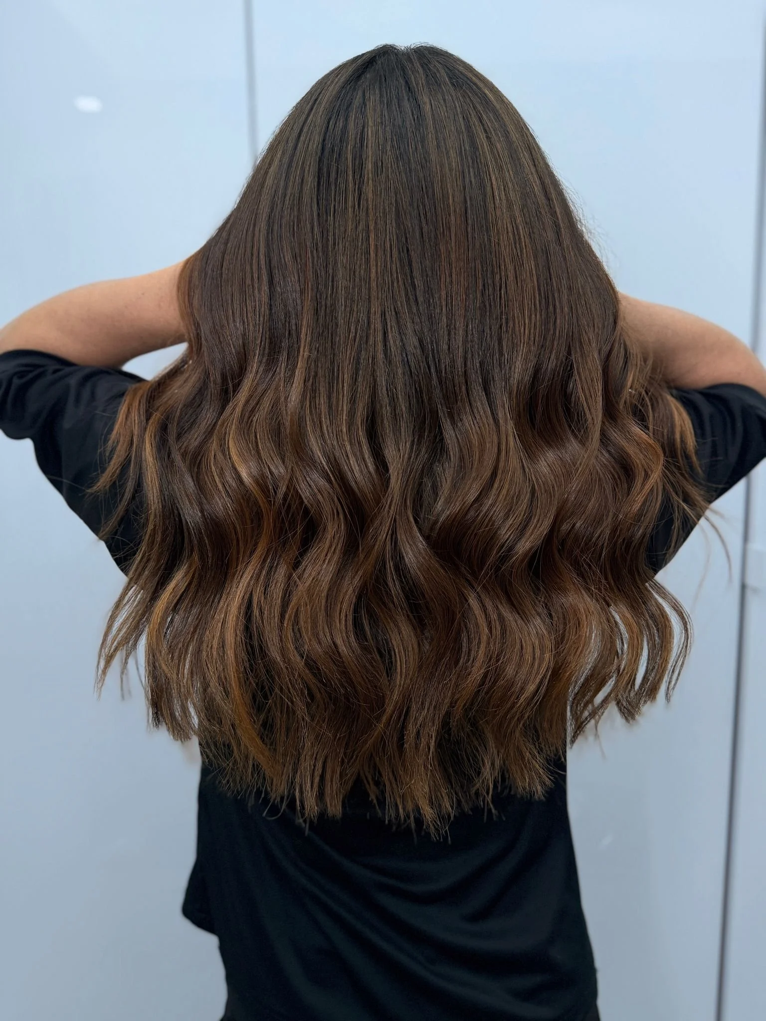 A person with long, wavy, brown hair with lighter highlights, wearing a black shirt, standing with their back to the camera, arms raised behind their head, against a plain light gray background.