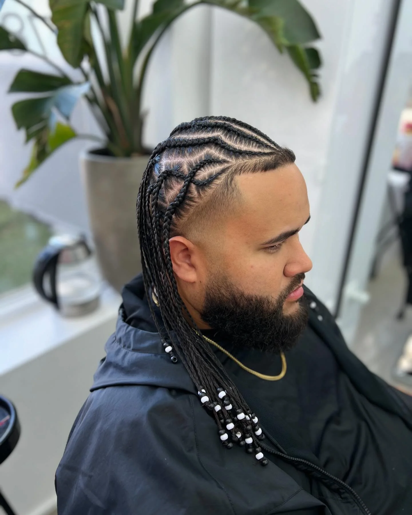 A man with braided hairstyle and a beard, wearing a black jacket and gold chain, sitting indoors near a large potted plant.