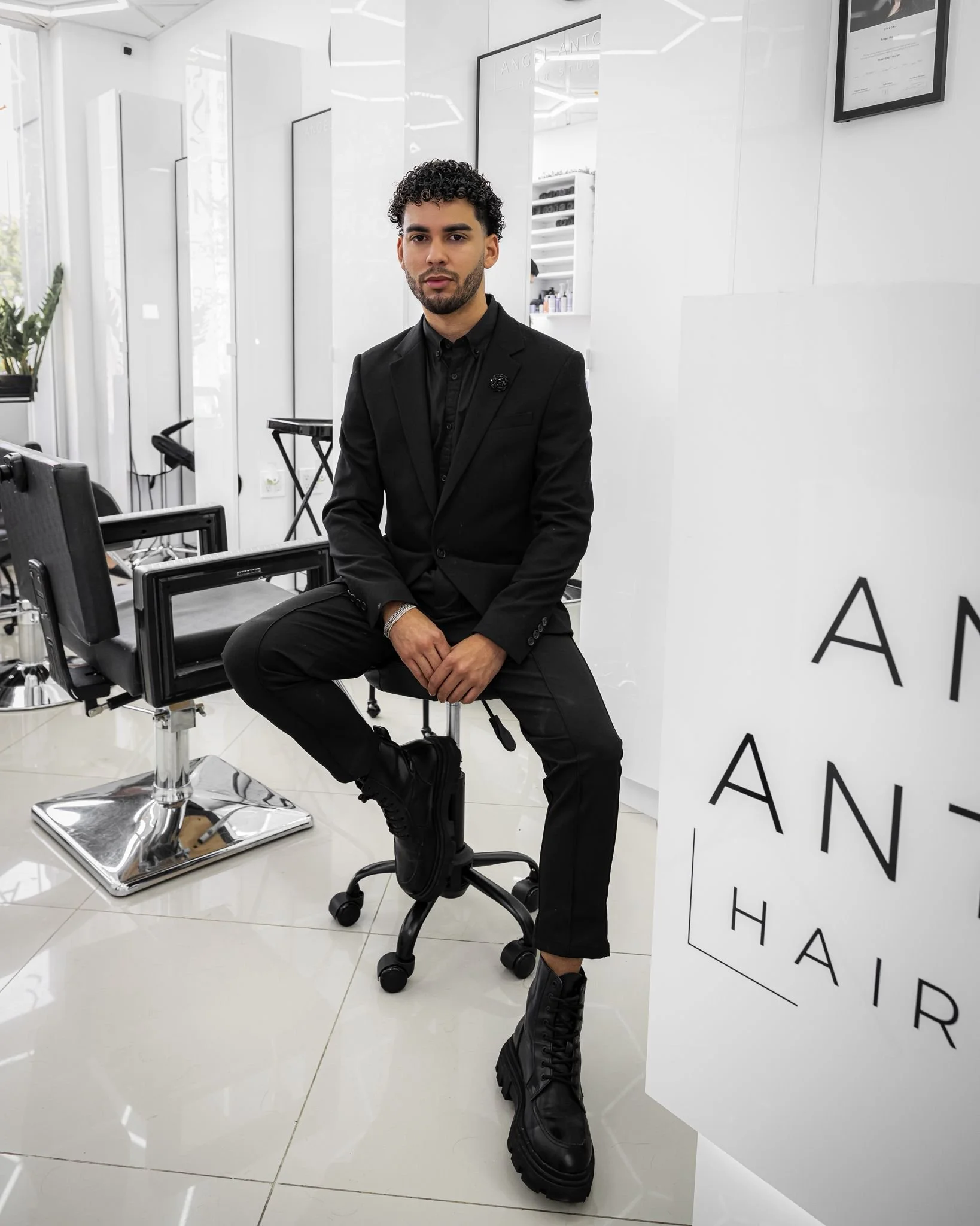 A man with curly hair and a beard, dressed in a black suit, sitting on a black office chair inside a modern, white-themed room, with black boots and a white wall with partially visible black text.