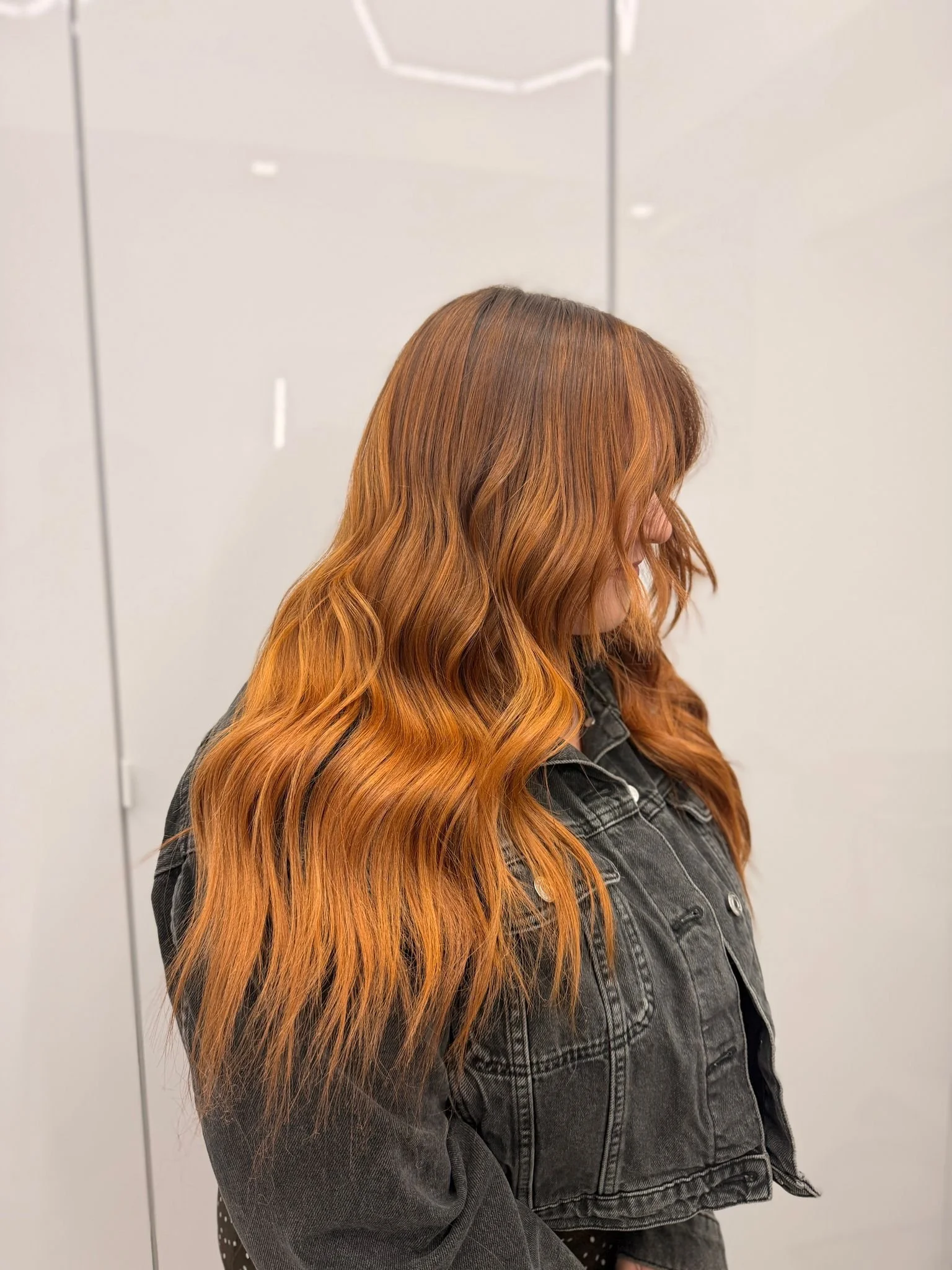 Side view of a woman with long wavy hair colored in shades of copper and auburn, wearing a black denim jacket with metal buttons, standing against a plain beige wall.