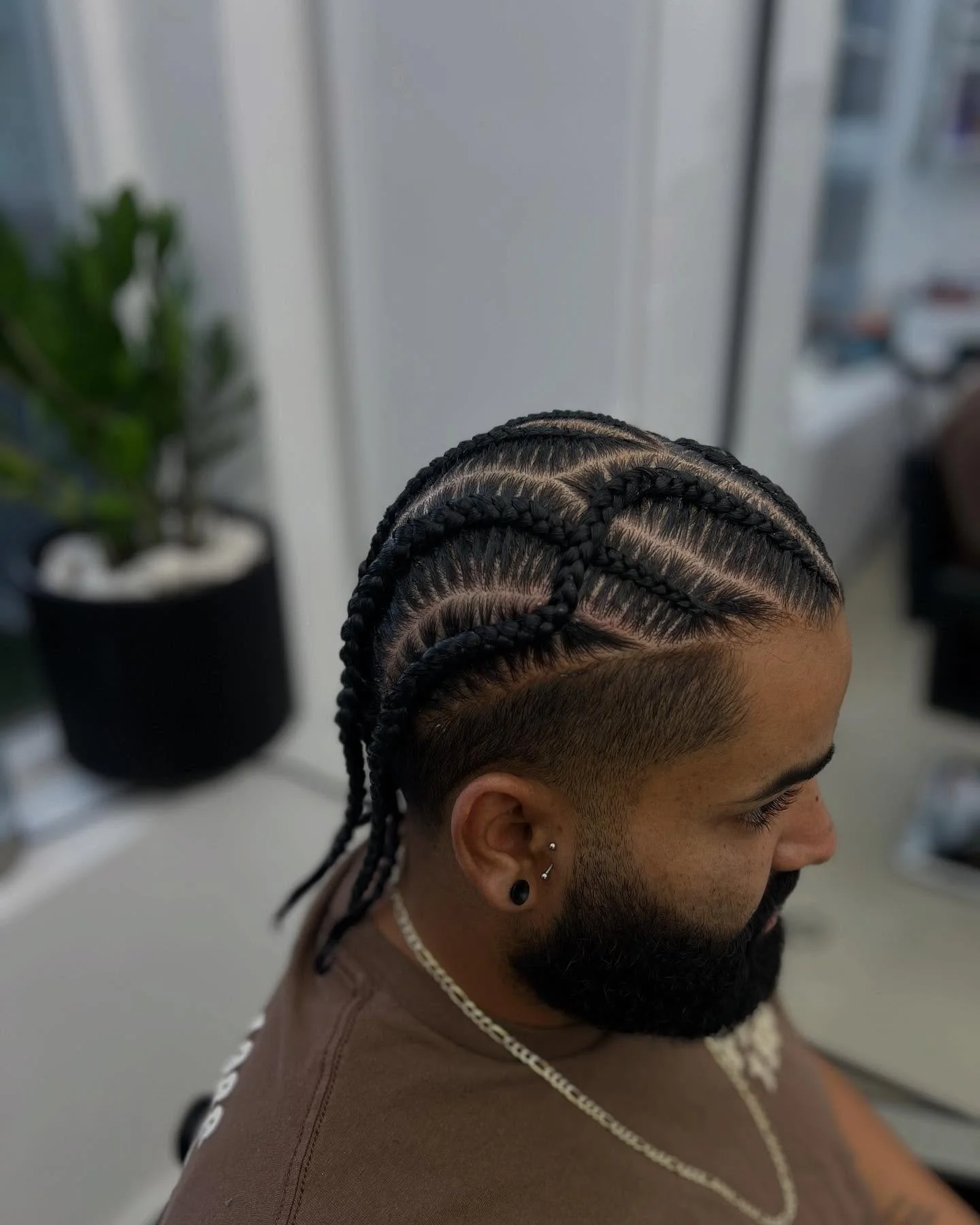 A man with braided cornrows hairstyle, a beard, and earrings, sitting in a salon with a blurred plant and window in the background.