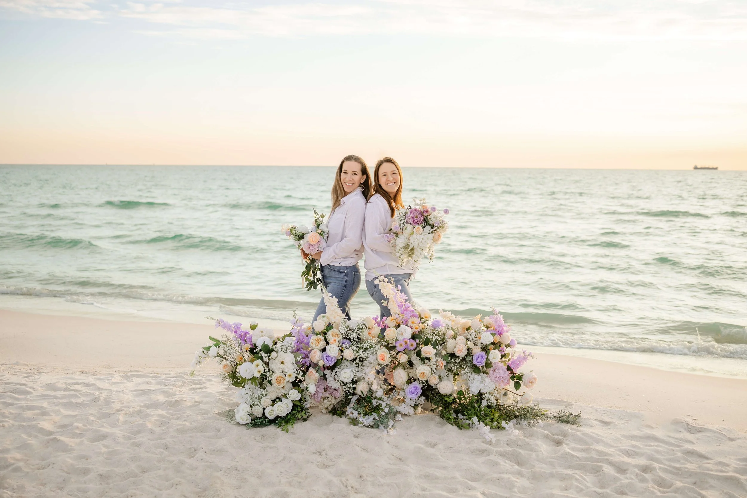 owners of Coastal Bloom standing back-to-back on a beach at sunset, holding bouquets of flowers, with luxury florals at Panama City beach florida.