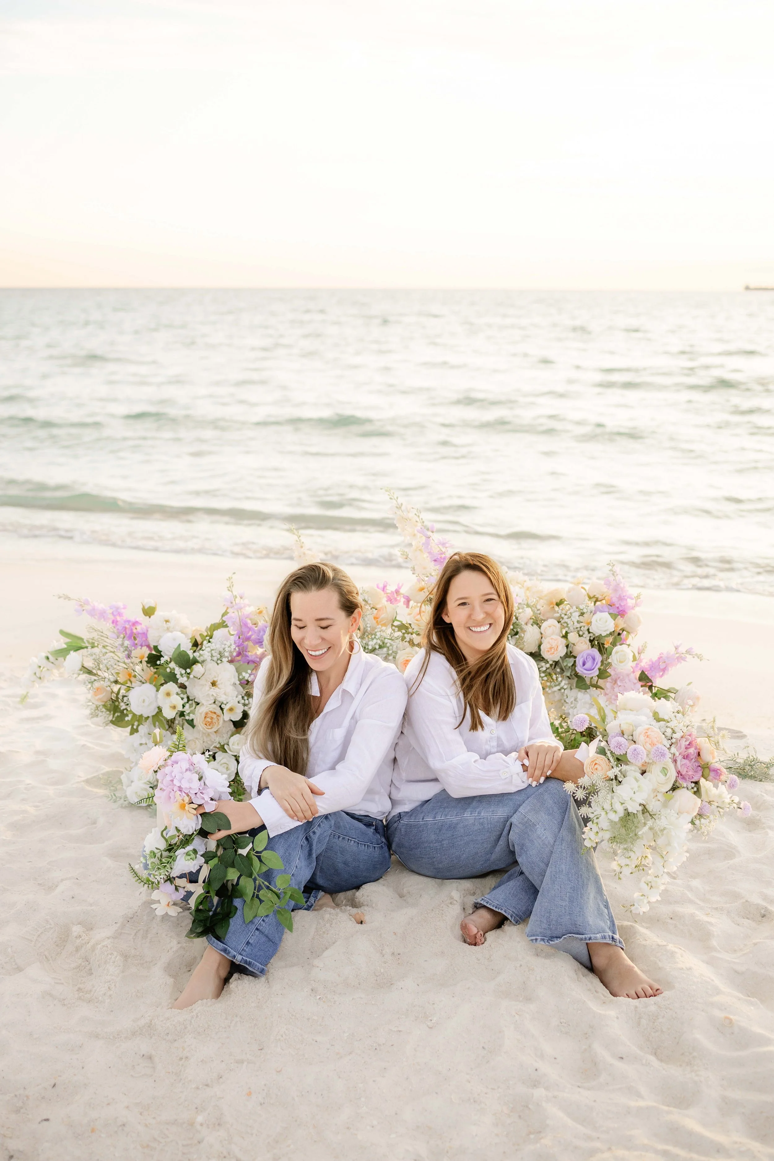 Owners of Coastal bloom sitting on a sandy beach with large floral arrangements behind them, in front of the ocean during sunset, in 30A Florida.