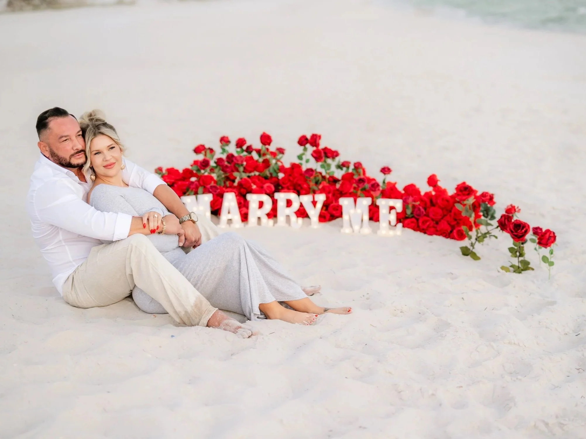 A couple sitting on the sand at the beach with a display of roses and large letters spelling 'Marry Me' in the background.