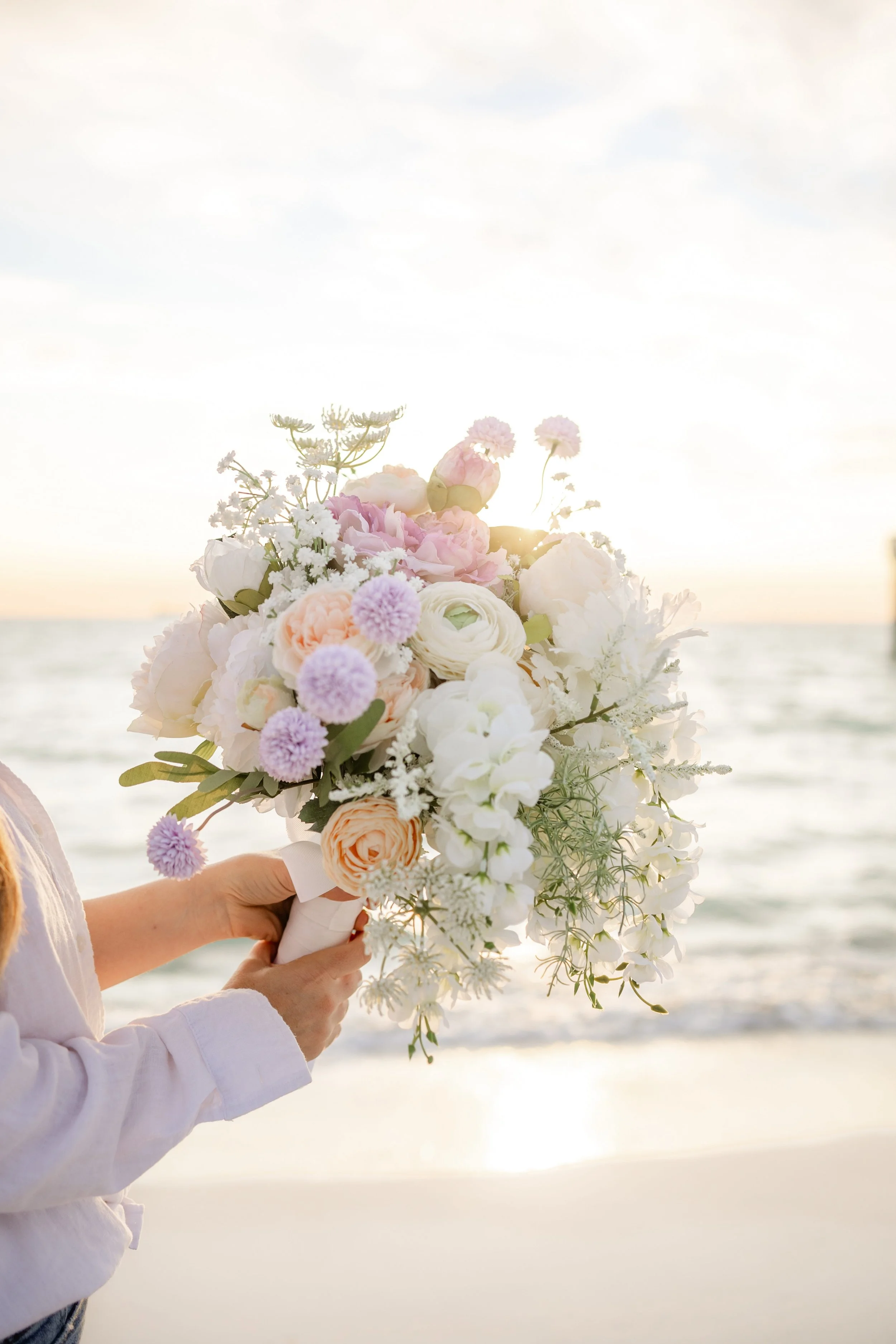 Person holding a white, pink, and purple bouquet of flowers on a beach at sunset in PCB.