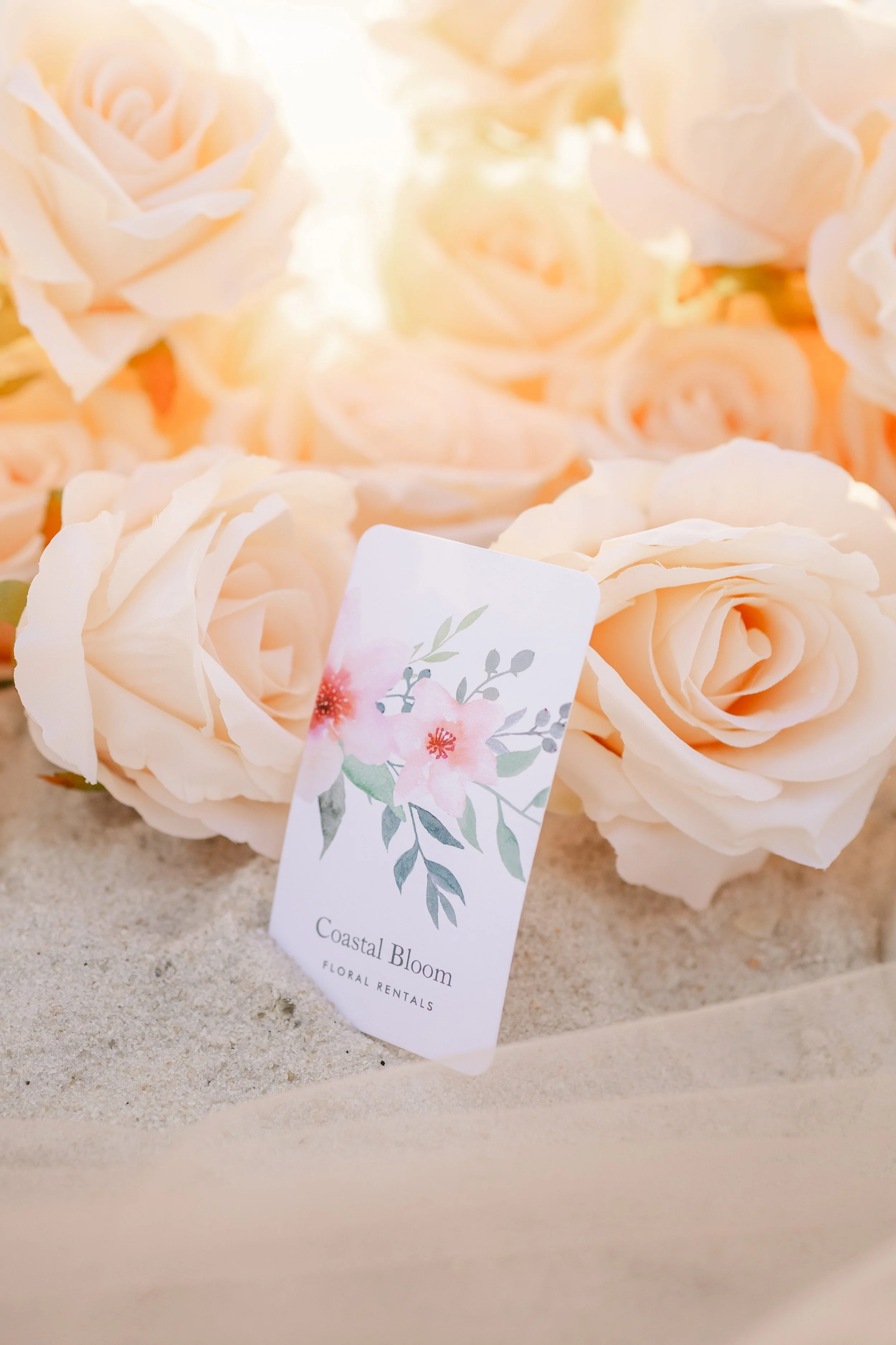 Cream-colored roses with a floral rental tag that says 'Coastal Bloom' on a sandy surface at the beach in the panhandle of florida.