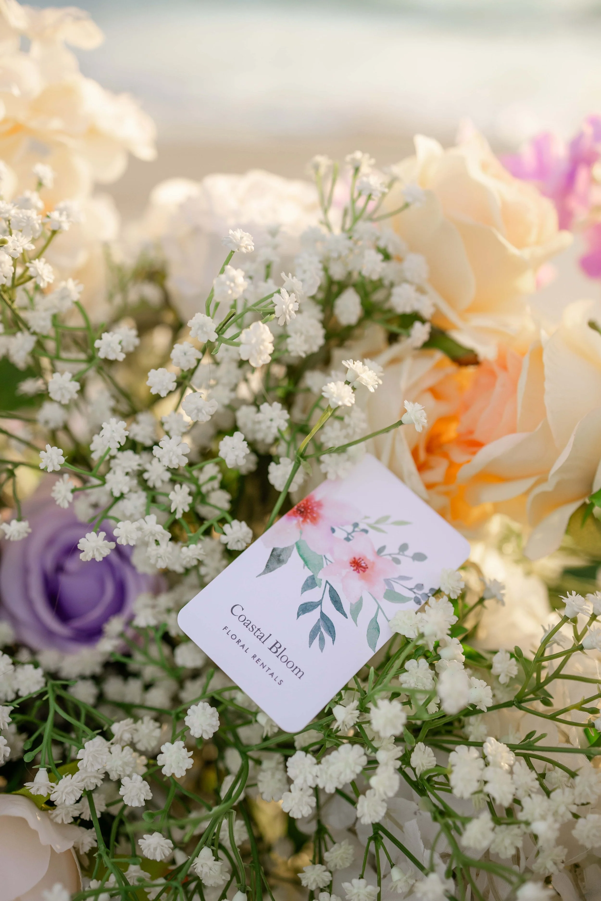 A bouquet of flowers with a card that reads "Coastal Bloom Floral Rentals." The bouquet includes white baby's breath, cream roses, and purple roses, with a softly blurred background in Panhandle of florida.