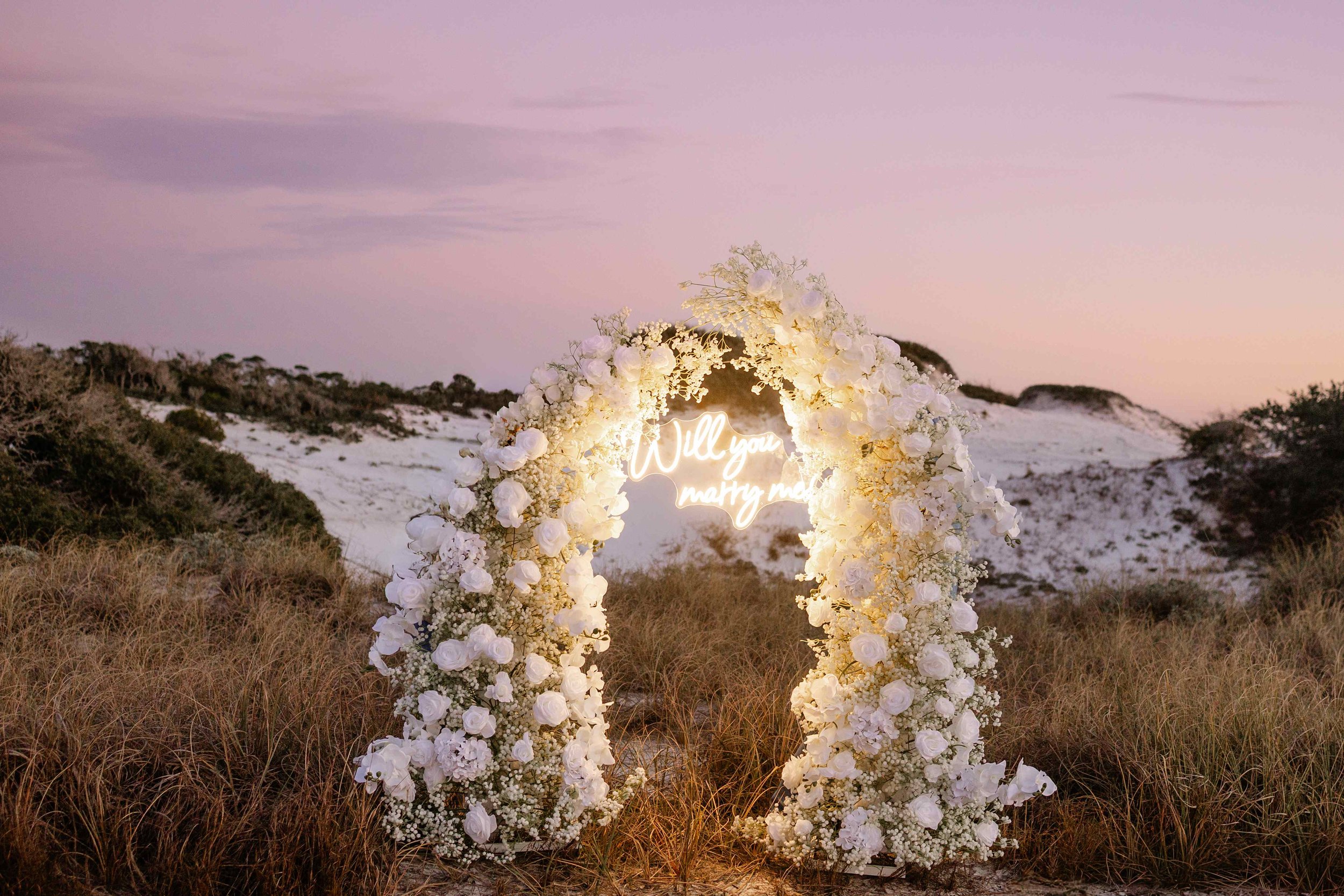 A floral wedding arch decorated with white roses and baby's breath, illuminated by a neon sign reading 'Will you marry me,' set against a beach landscape at sunset in Panama City beach Florida/ 30a.