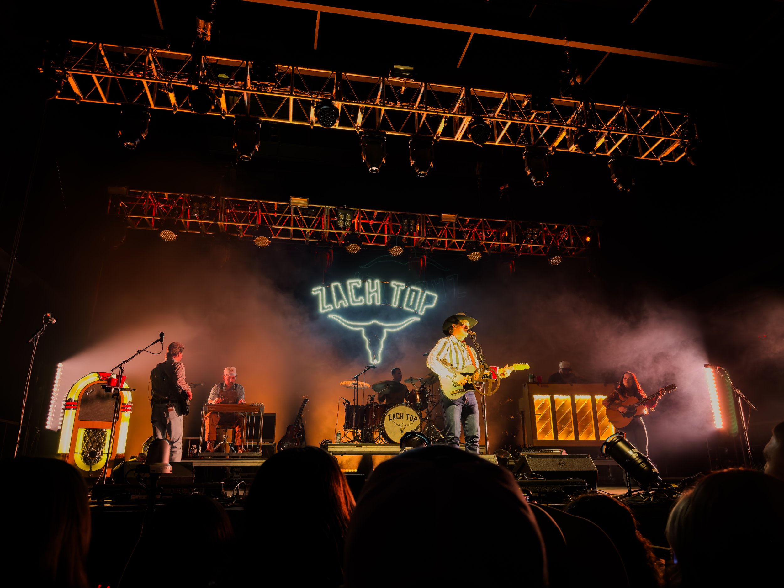 Live concert performance with a band on stage, featuring a singer in a cowboy hat, orange lighting, and a foggy atmosphere, with the words 'ZACH TOP' in neon lights.