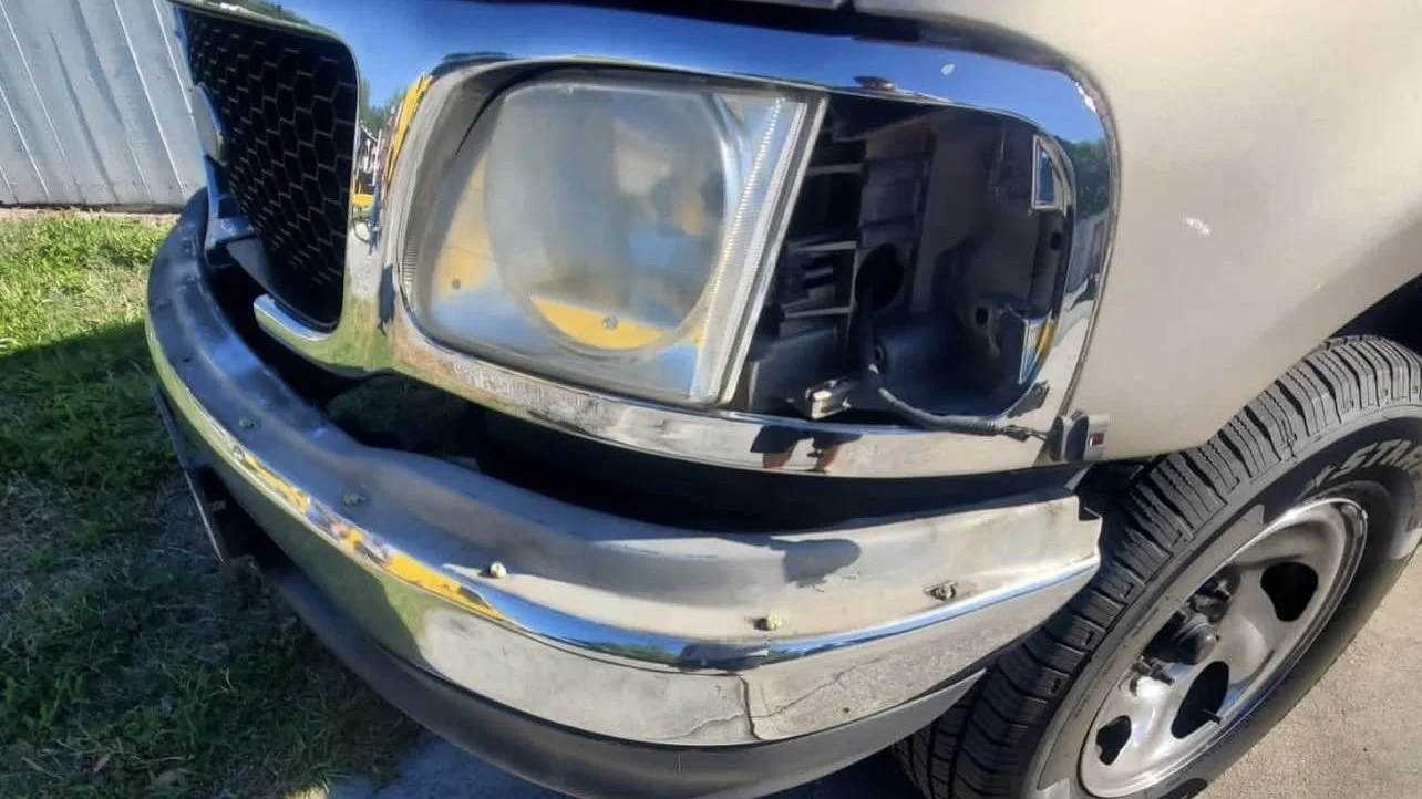 Front view of a beige pickup truck with a damaged headlight and missing grille, showing a chrome bumper and tire.