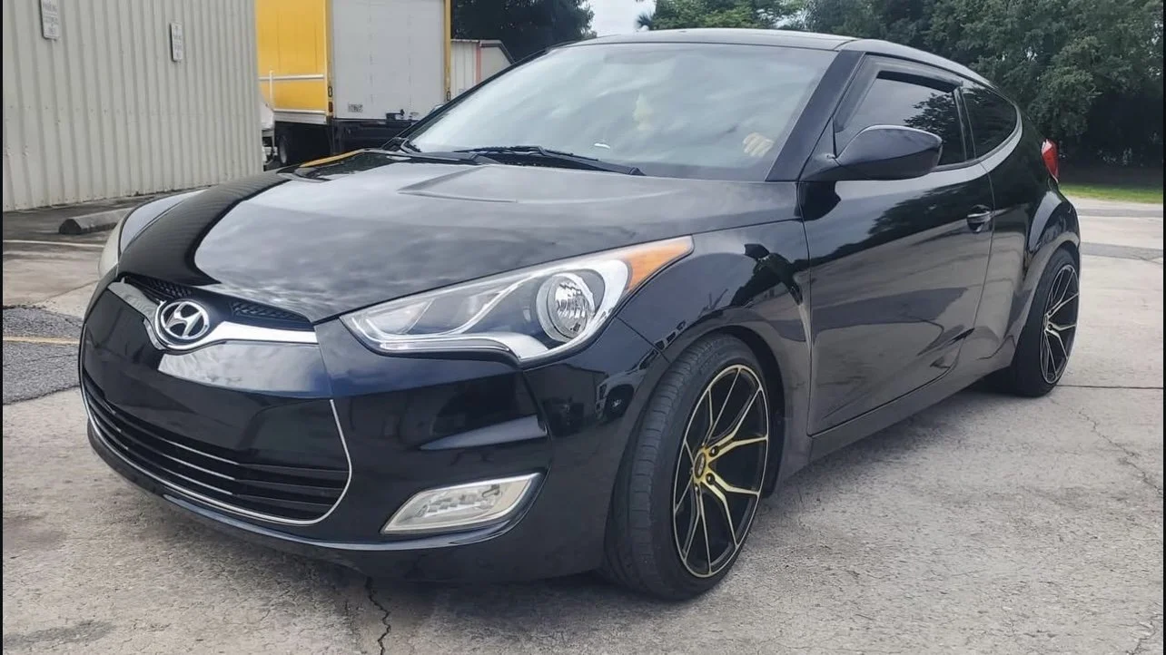 Black Hyundai Veloster with custom black and gold wheels parked outdoors with a building and tree in the background.