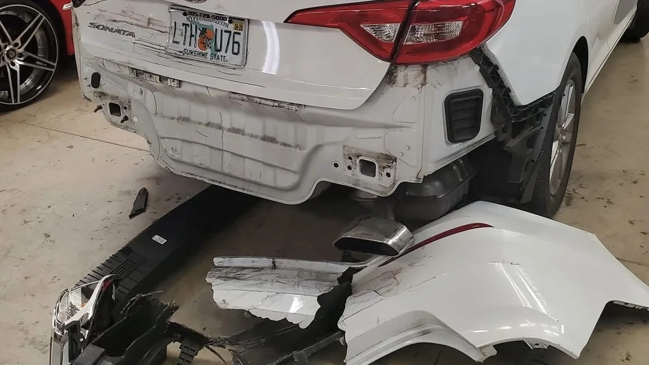 A rear-end collision damage on a white car with detached bumper and broken tail light on the ground.
