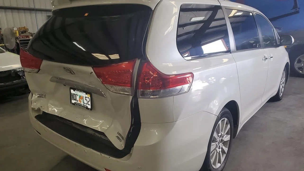 White minivan with damaged rear bumper and dented tailgate inside a garage.