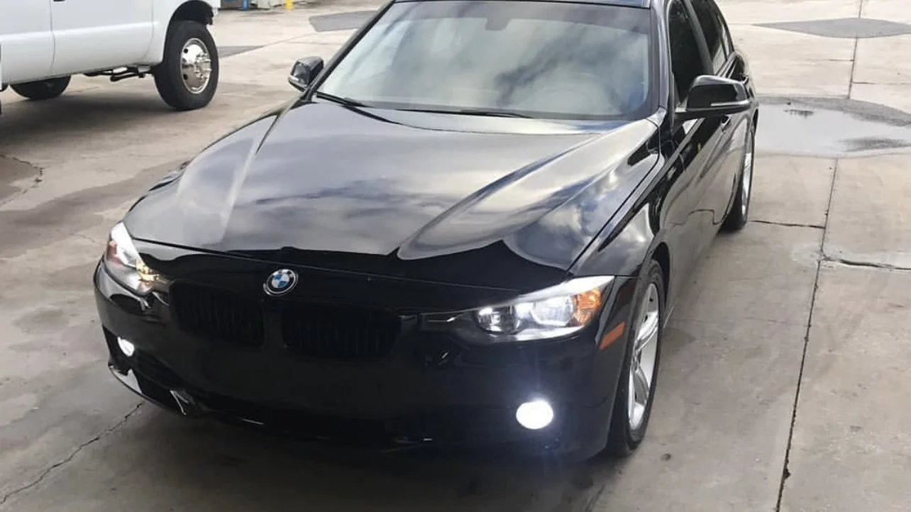 Black BMW sedan parked on pavement, with headlights on. A white truck is partially visible in the background.