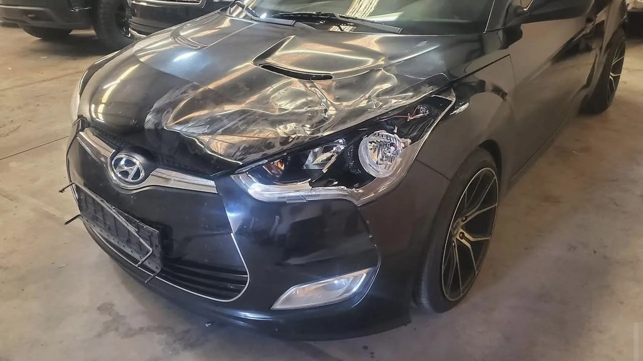 Front view of a damaged Hyundai car with a dented hood and headlight in a garage.