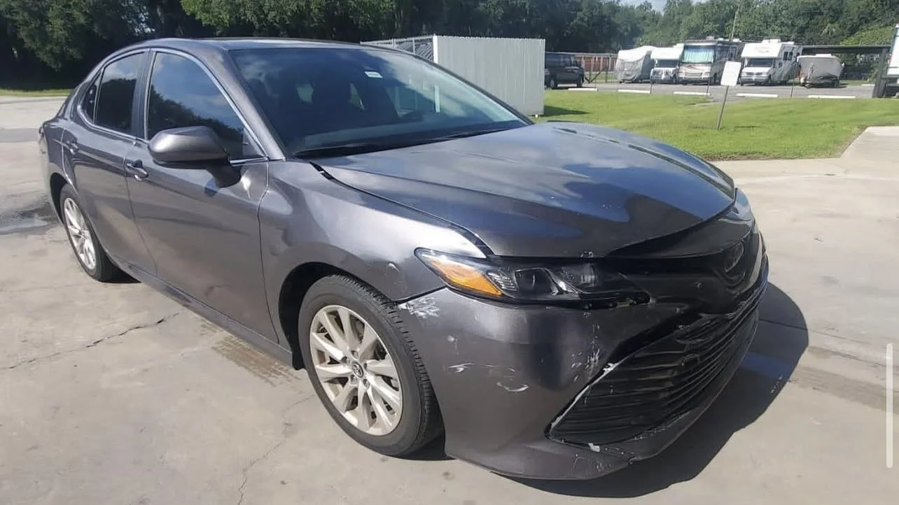 Damaged gray sedan with visible front bumper damage, parked on a concrete surface in an outdoor area with grass and trees in the background.