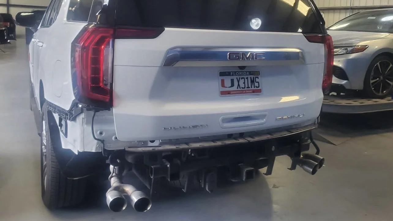 Rear view of a white GMC vehicle in a garage, showing a Florida license plate and a missing rear bumper revealing exhaust pipes.