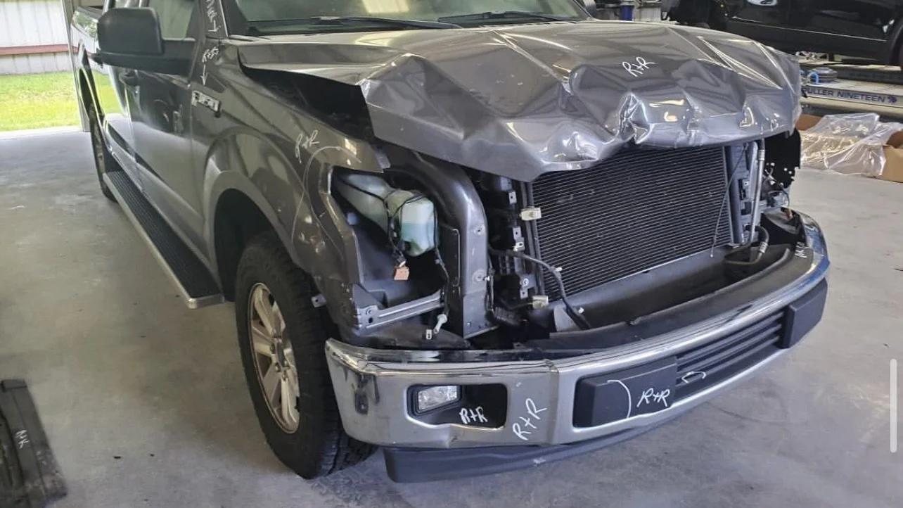 Damaged gray pickup truck with crumpled hood and exposed front assembly in a garage.