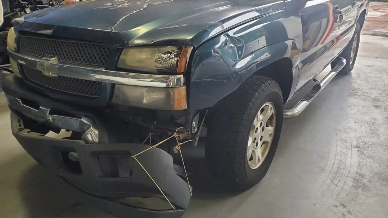 Damaged blue pickup truck with a broken front bumper and dented fender in a garage.