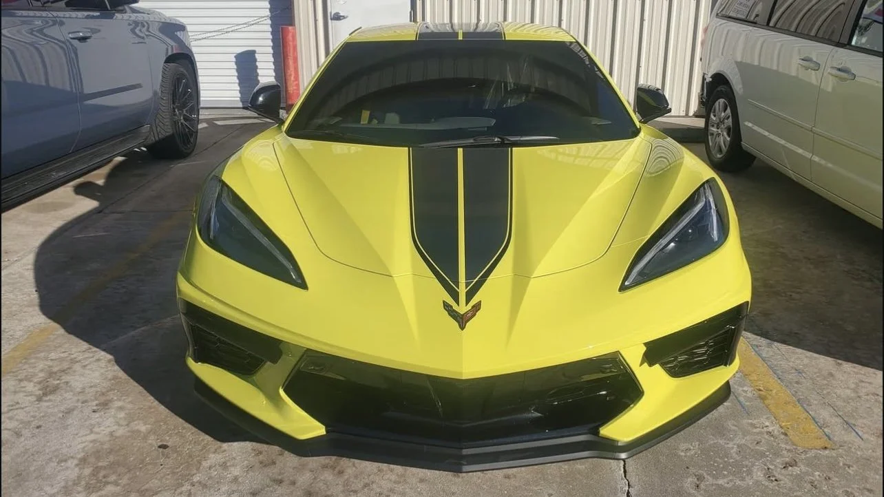 Front view of a yellow sports car with black racing stripes parked between two vehicles.