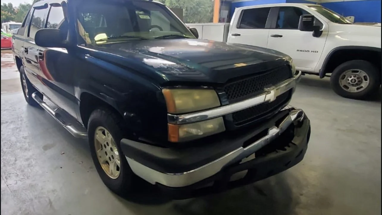 Black Chevrolet truck with front bumper damage inside a garage, parked next to a white pickup truck.
