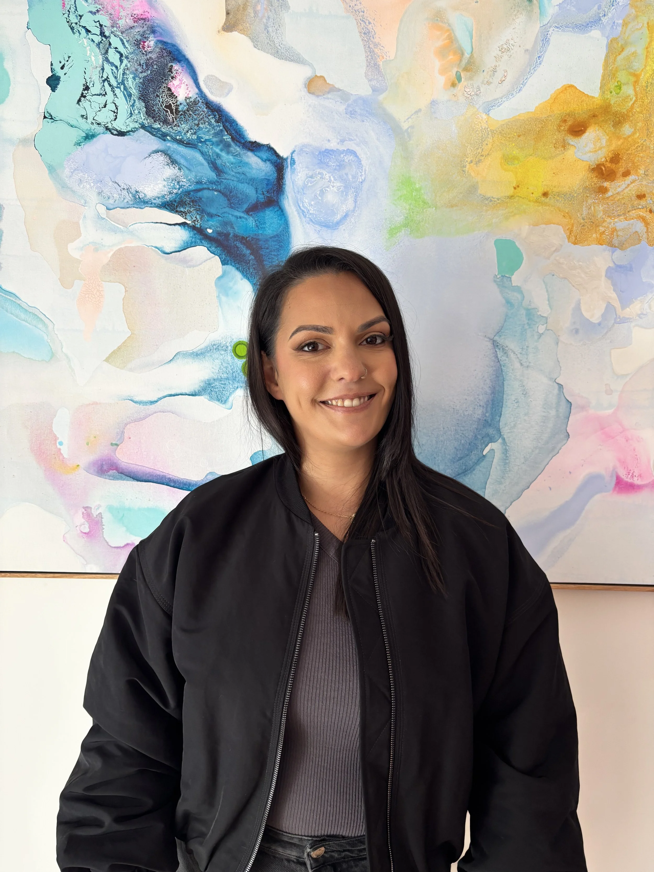 A woman with black hair wearing a black jacket and gray shirt smiling in front of an abstract colorful painting.