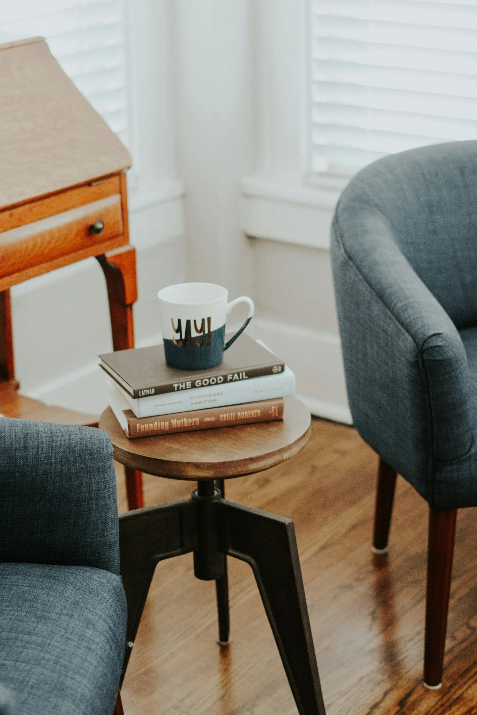 A cozy living room with a small round wooden table holding a book and a mug, between two upholstered chairs, with a wooden side table and white shuttered windows in the background.