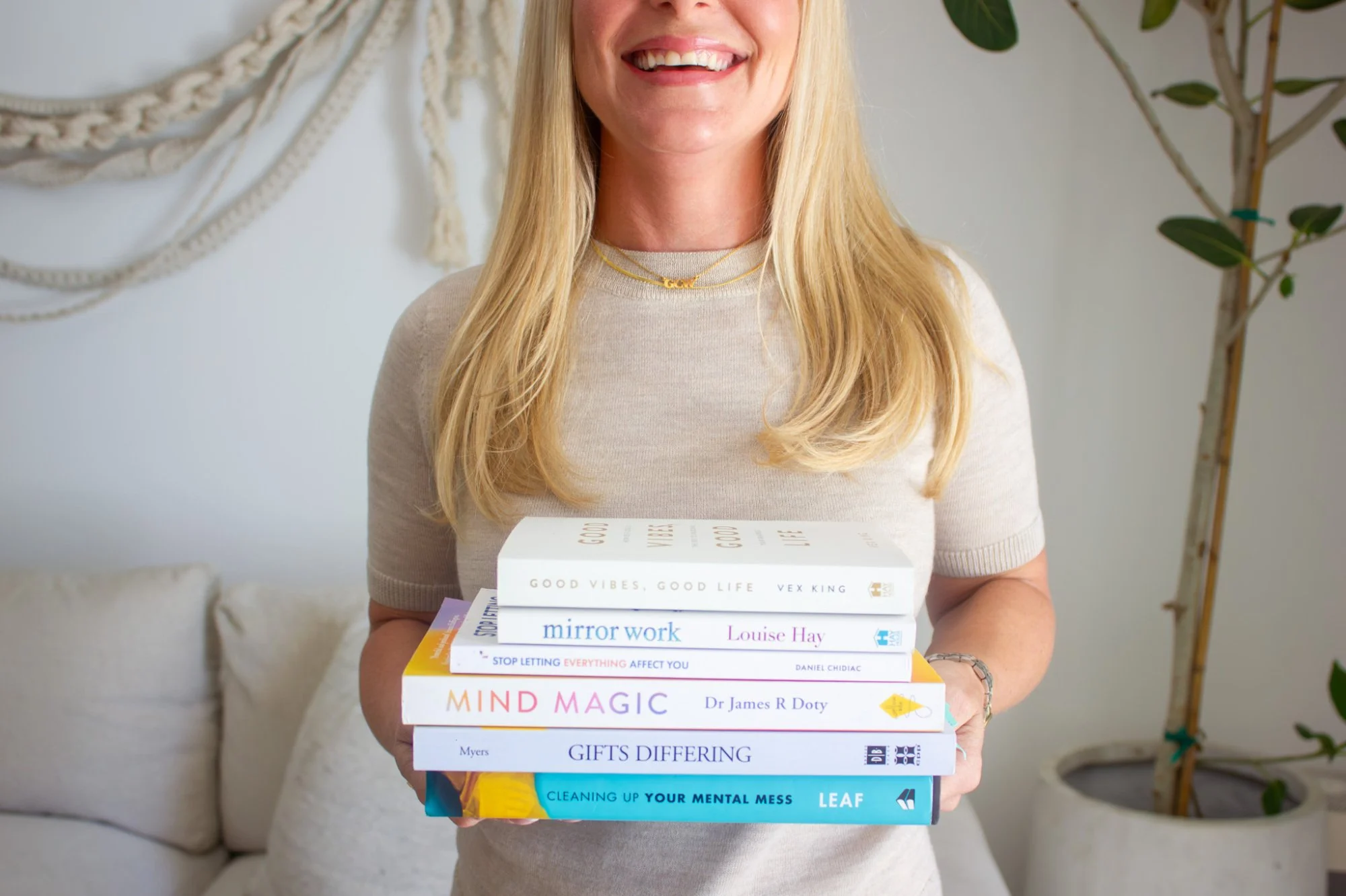 A woman with long blonde hair smiling, wearing a beige top and gold jewelry, holding a stack of books.