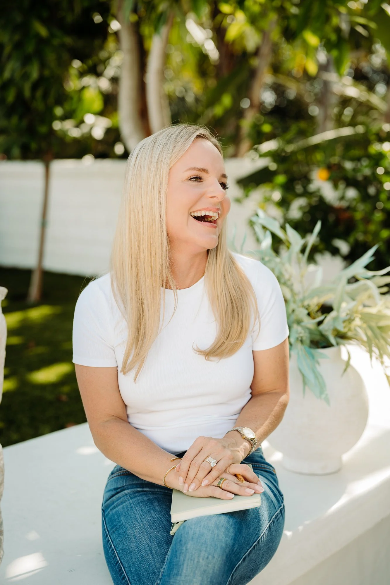 A woman with long blonde hair, wearing a white T-shirt and blue jeans, sitting outdoors with a smile on her face. She is accessorized with rings, a bracelet, and a watch, and is holding a notebook on her lap. The background features lush green trees and plants.