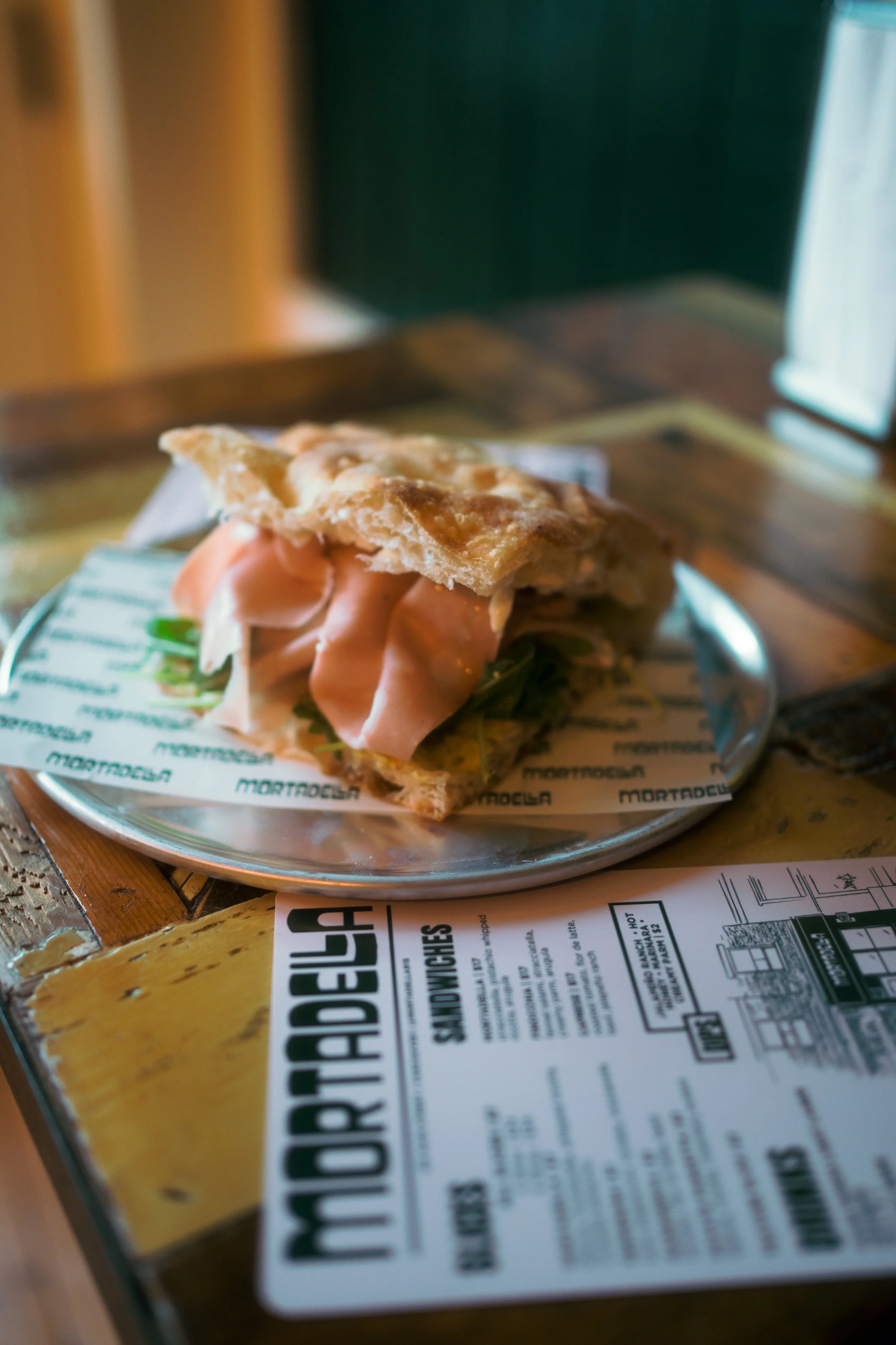 A sandwich with slices of ham and greens on a rustic wooden table, with a menu or flyer partially visible underneath.