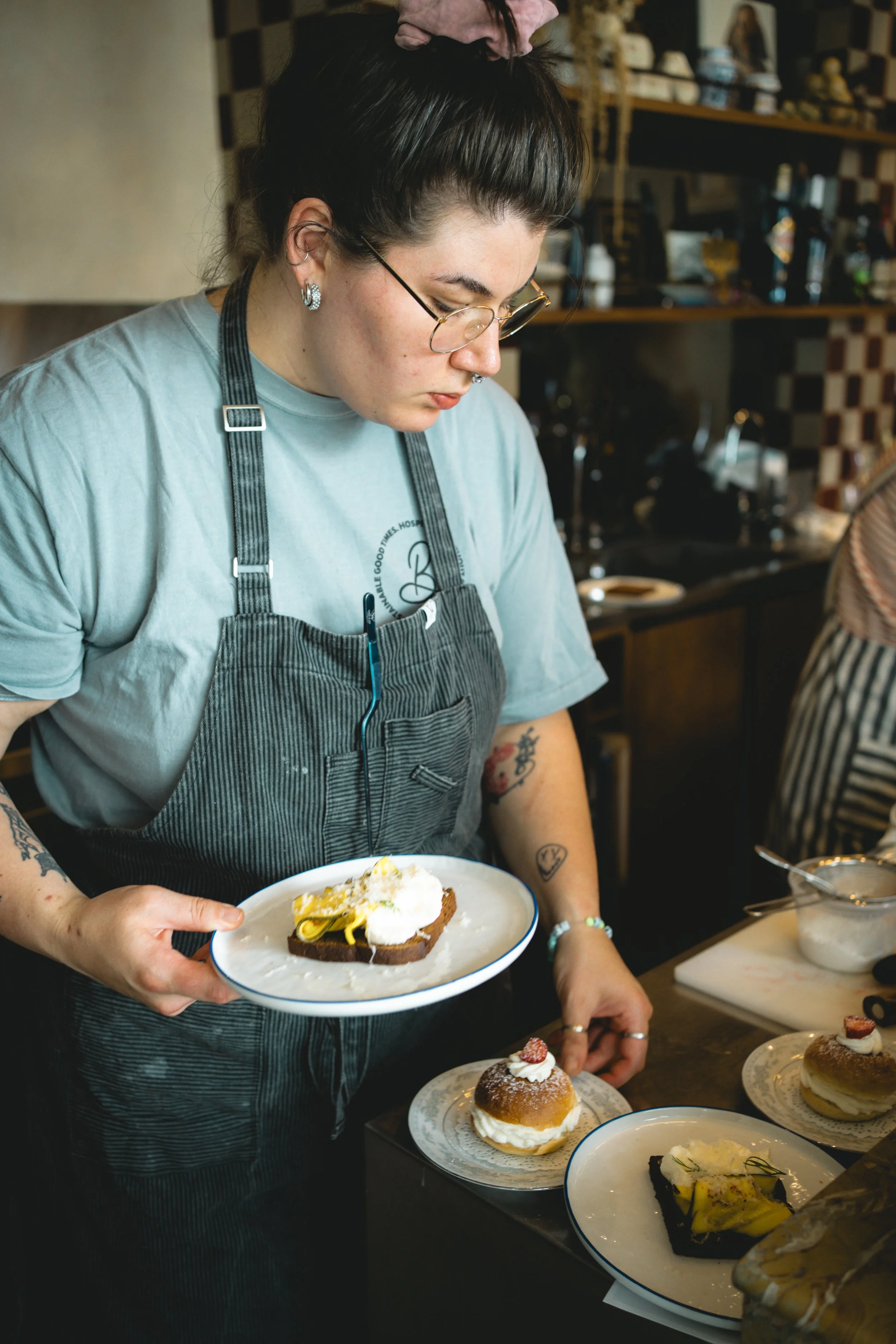 A woman wearing glasses, a grey t-shirt, and a striped apron is holding a plate with a slice of cake, surrounded by other desserts on the counter in a cozy bakery or cafe.