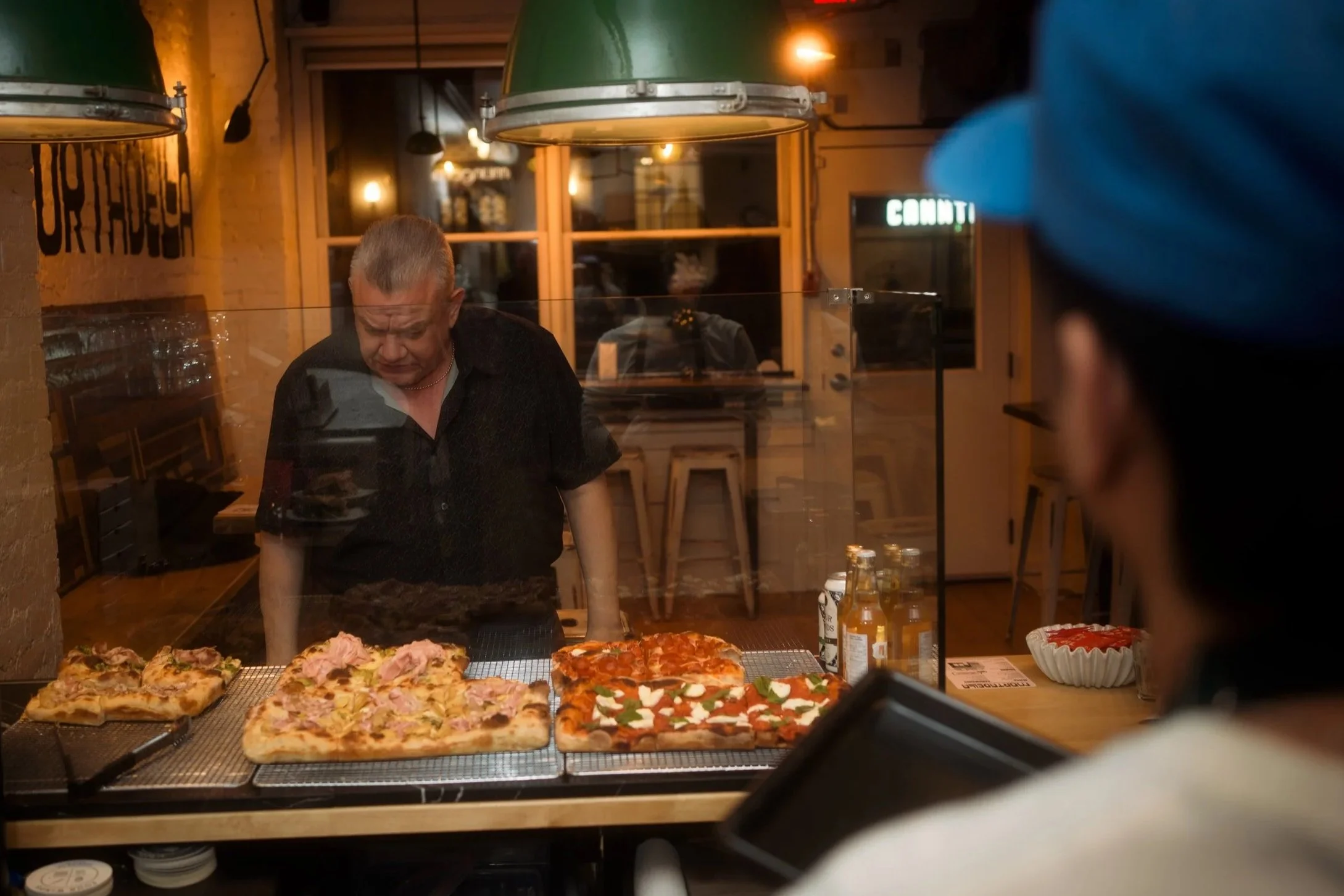 Person ordering pizza at a pizzeria counter with three pizzas on display behind a glass barrier.