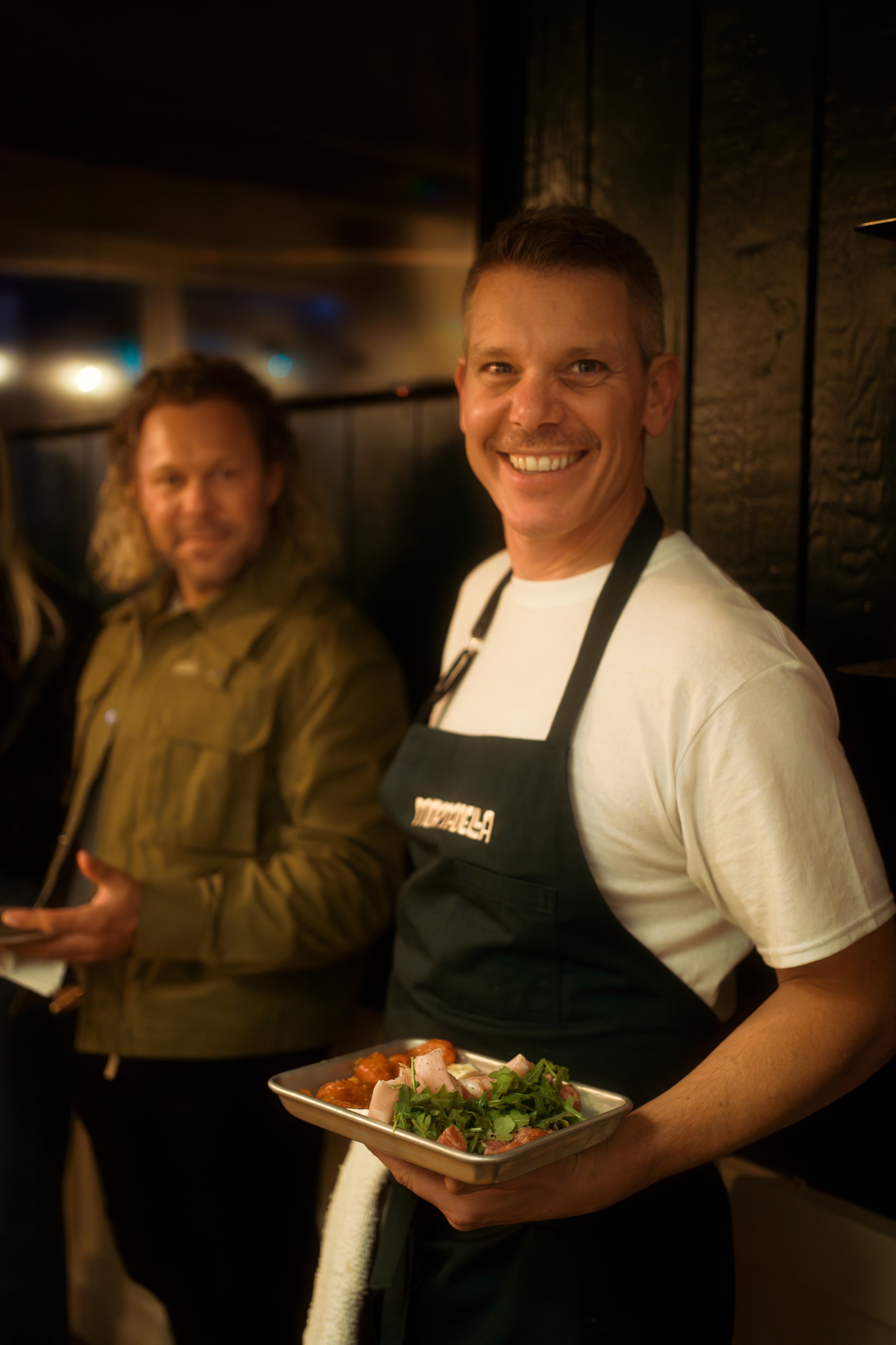A smiling man in a white t-shirt and black apron holding a tray of food, with another person blurred in the background in a dimly lit setting.
