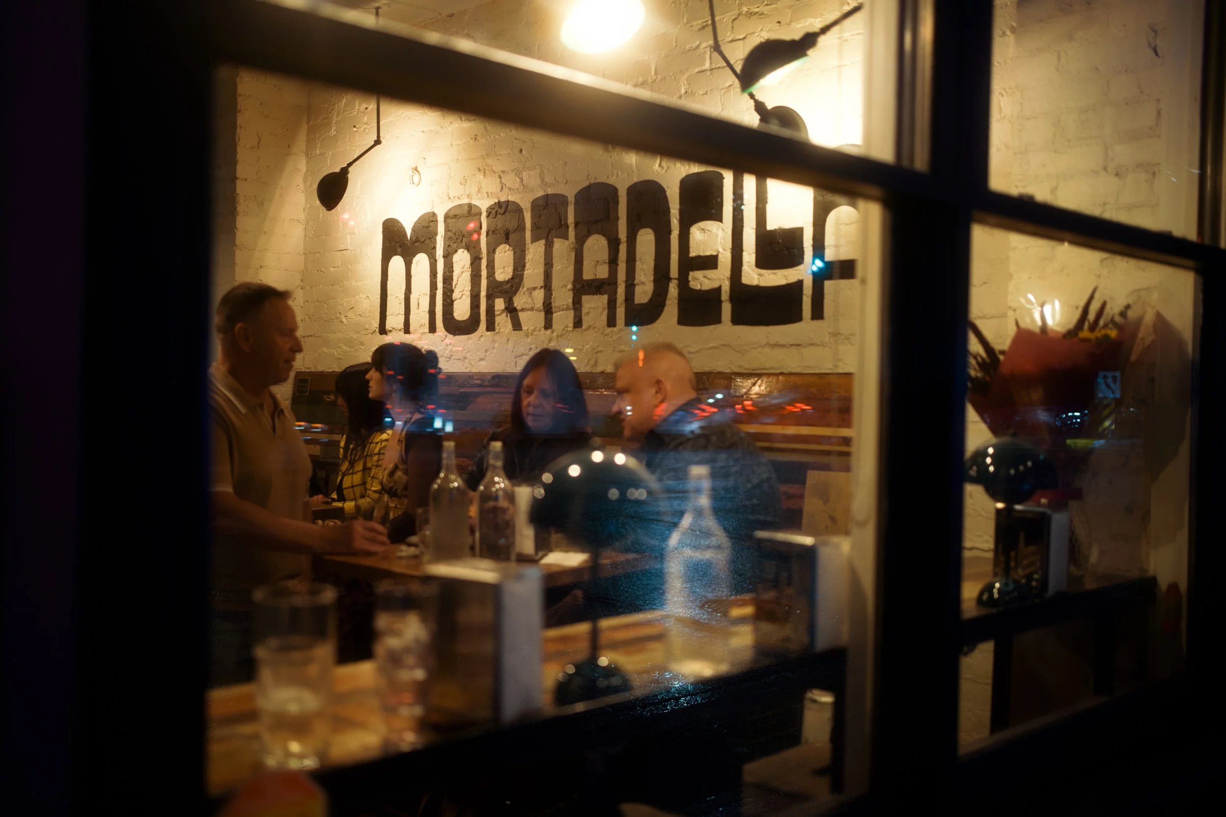 People sitting at a table inside a restaurant with a sign saying 'MORTADELLO' on the brick wall, seen through a window at night with reflections of city lights.