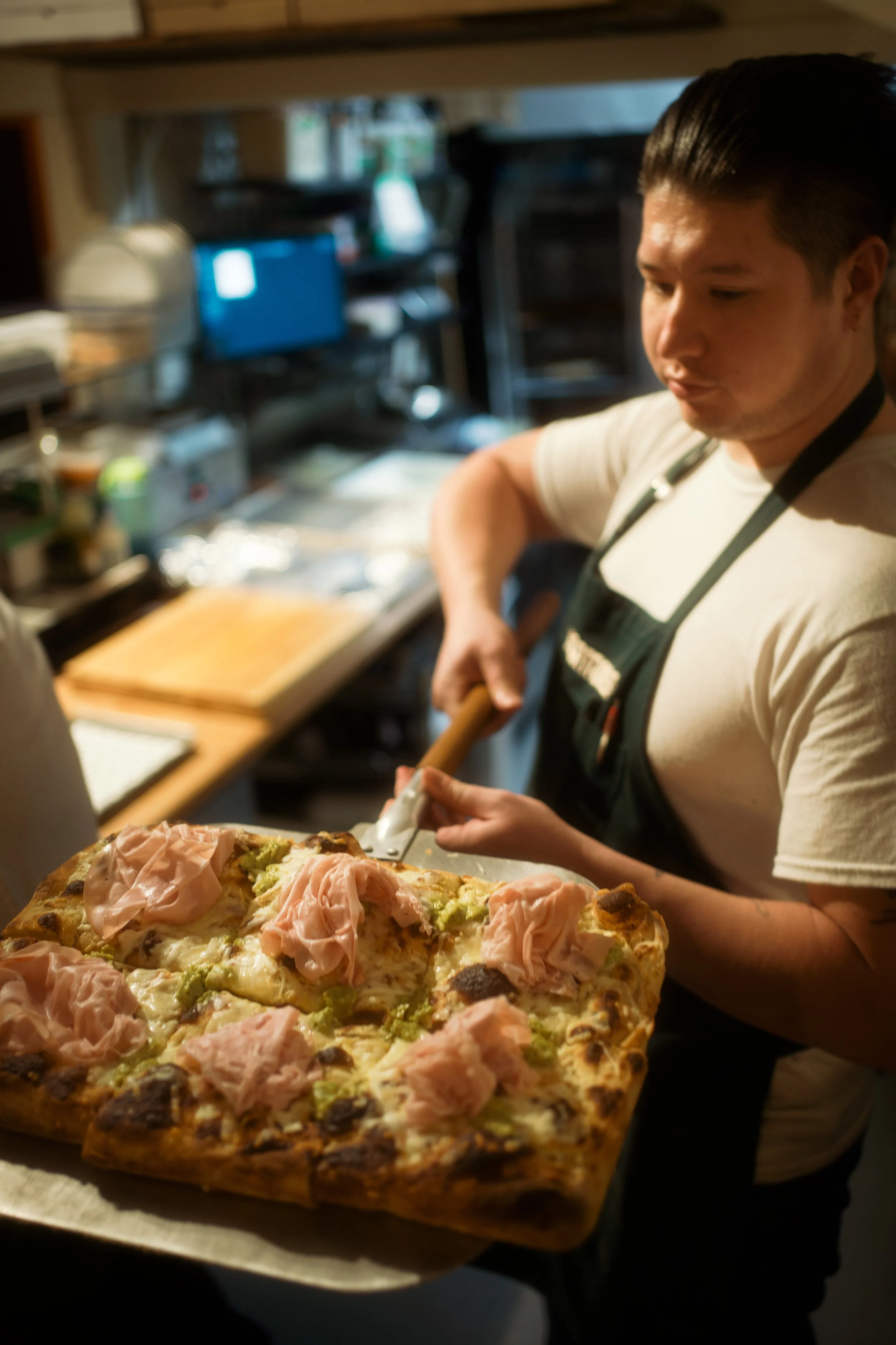 A person holding a freshly baked pizza topped with slices of ham and green vegetables.