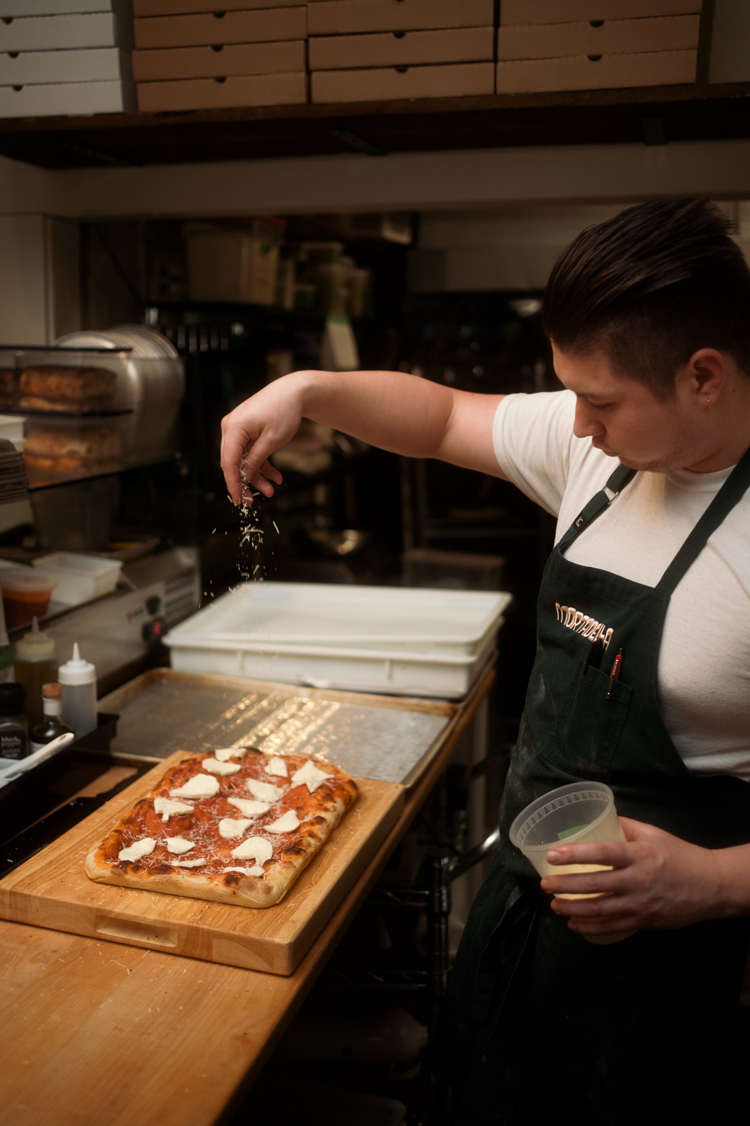 A person sprinkling grated cheese over a rectangular pizza topped with tomato sauce and cheese, on a wooden cutting board in a kitchen.