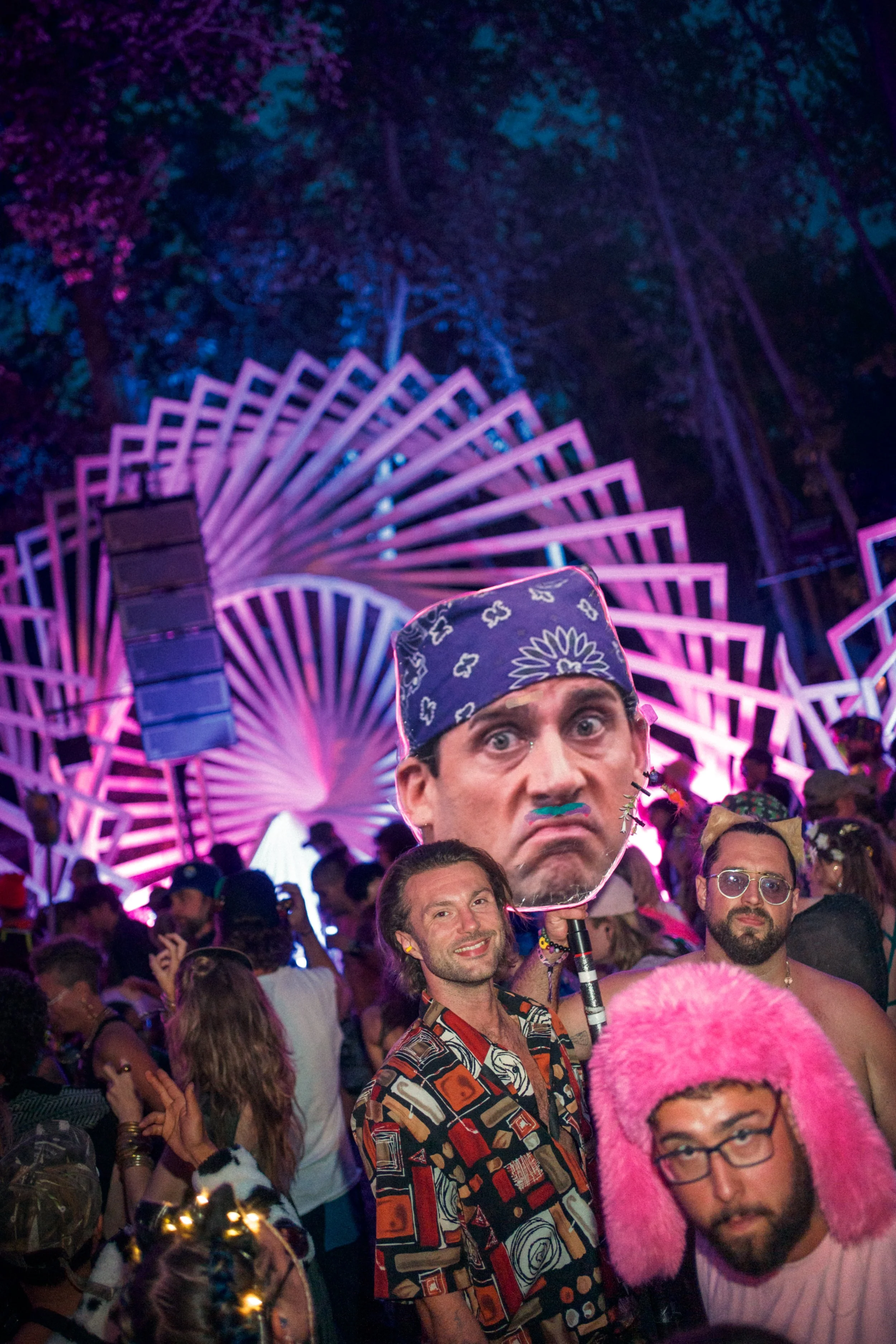 At a lively outdoor event with colorful lighting and a crowd of people in costume, one person holds a large handheld mask of a man's face with a stern expression, while others wear various playful accessories including a pink fluffy hat and sunglasse