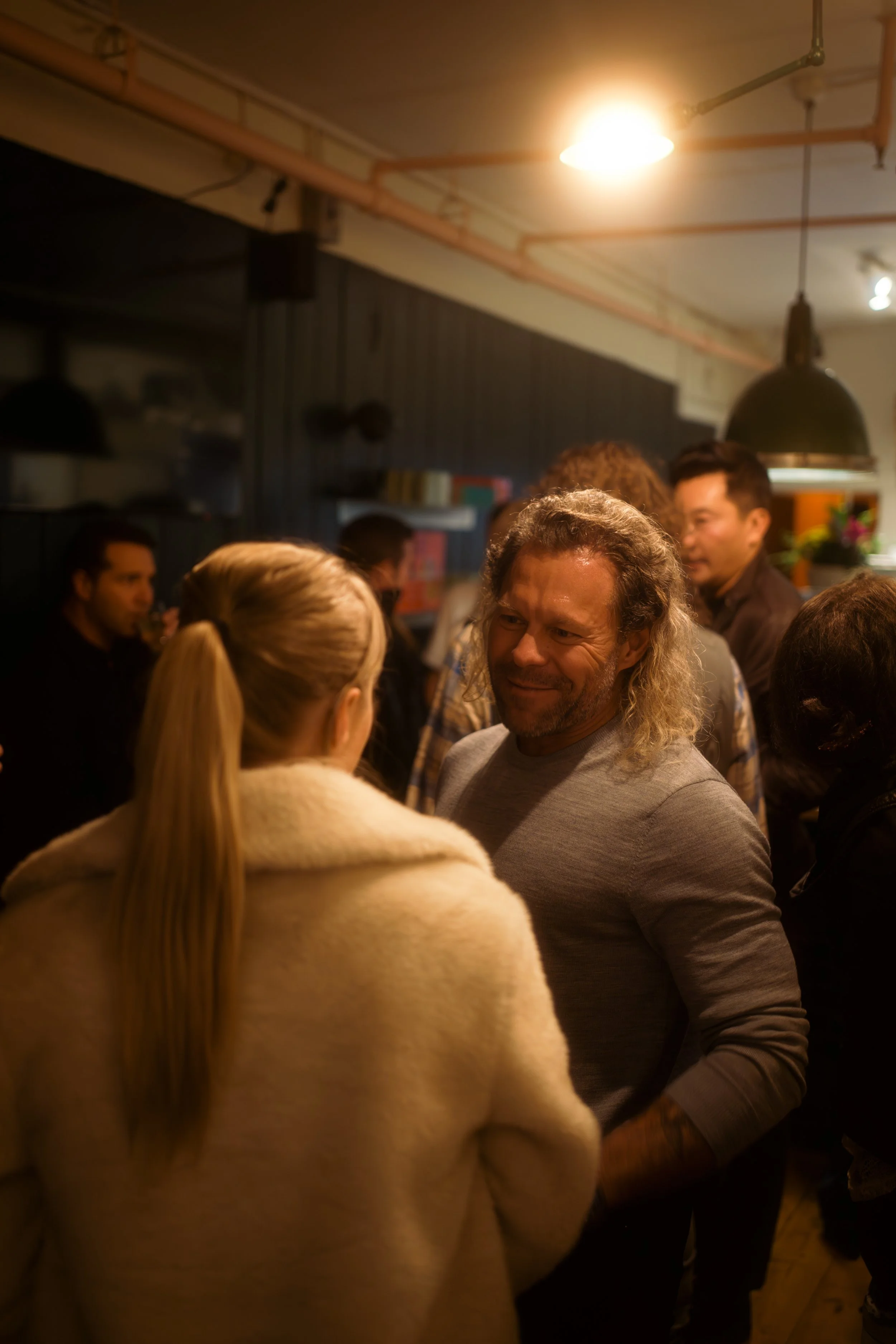 A social gathering indoors with people engaged in conversation, illuminated by warm lighting, featuring a man with long wavy hair and a woman with long blonde hair in the foreground.