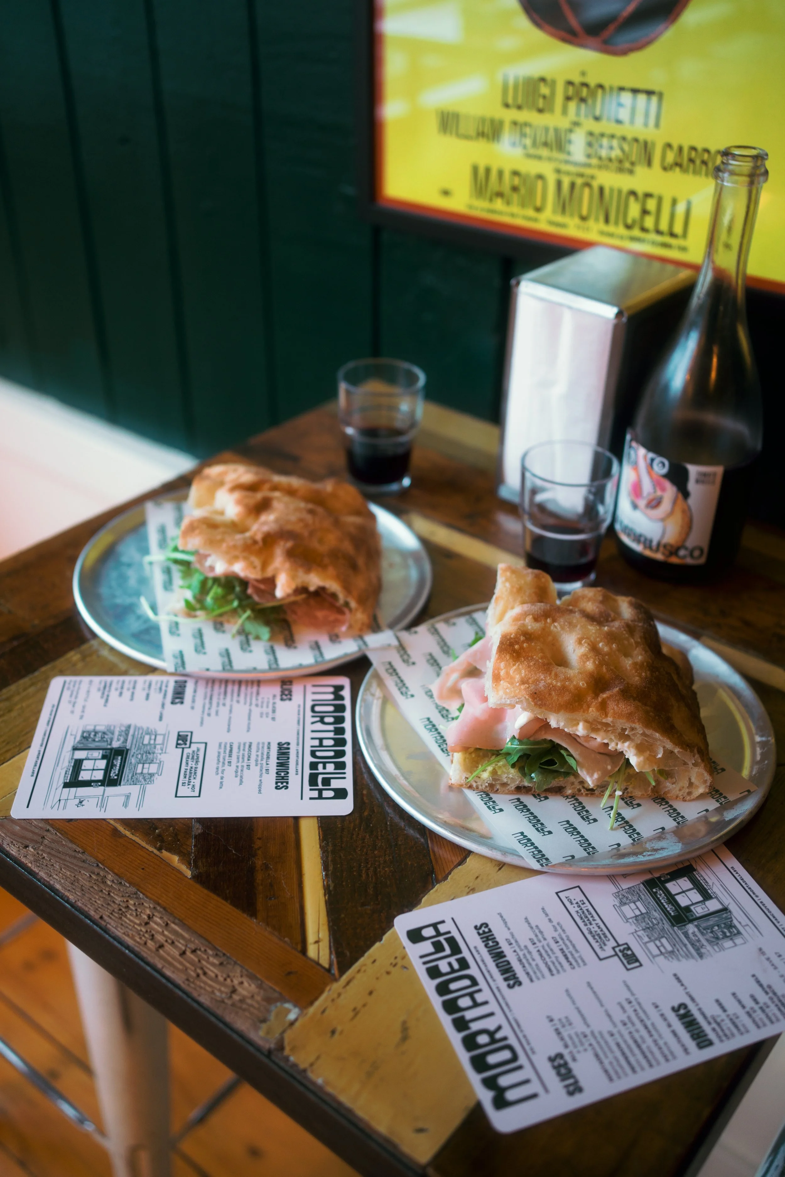 Two sandwiches with deli meats and greens on metal plates, on a wooden table with menus and another plate, two small glasses of dark beverage, and a bottle of sparkling water outside a restaurant with a yellow and black sign.