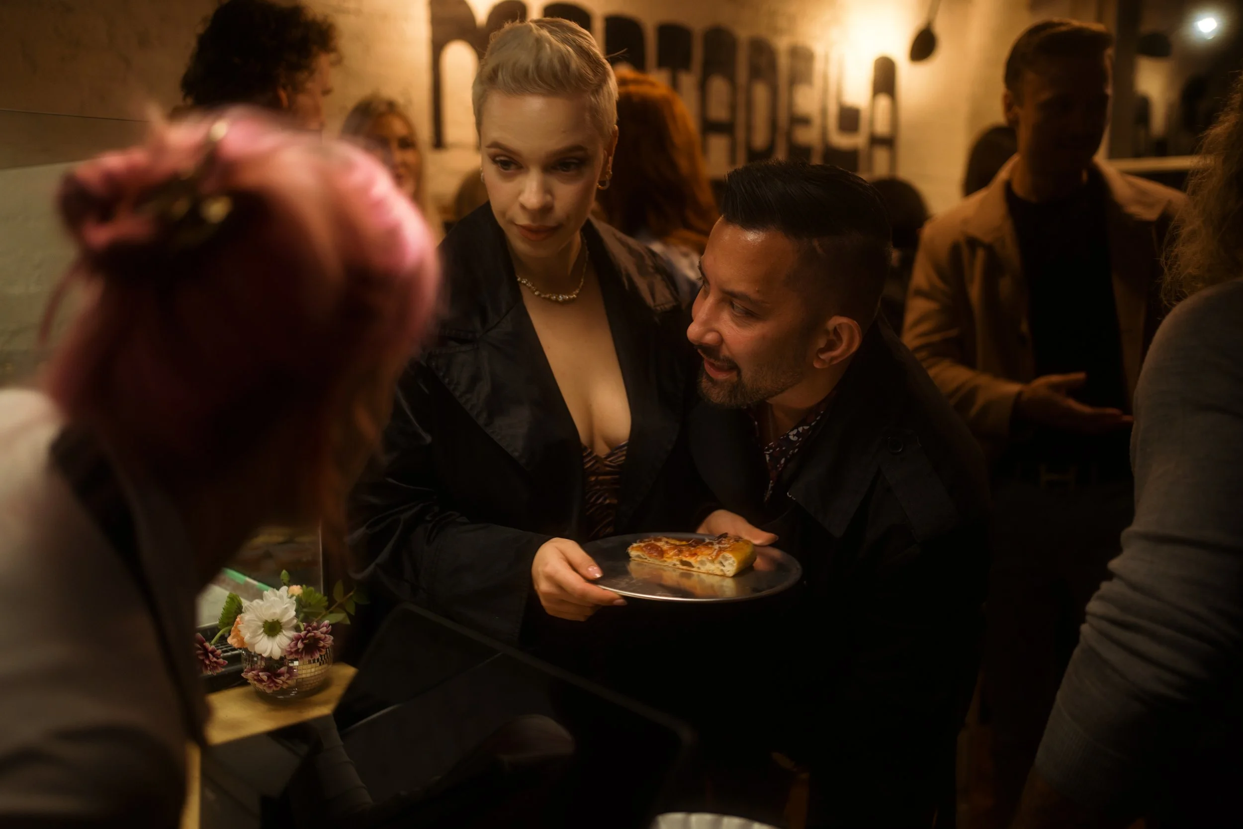 People socializing in a dimly lit restaurant, with a woman holding a pizza and talking to others around her.