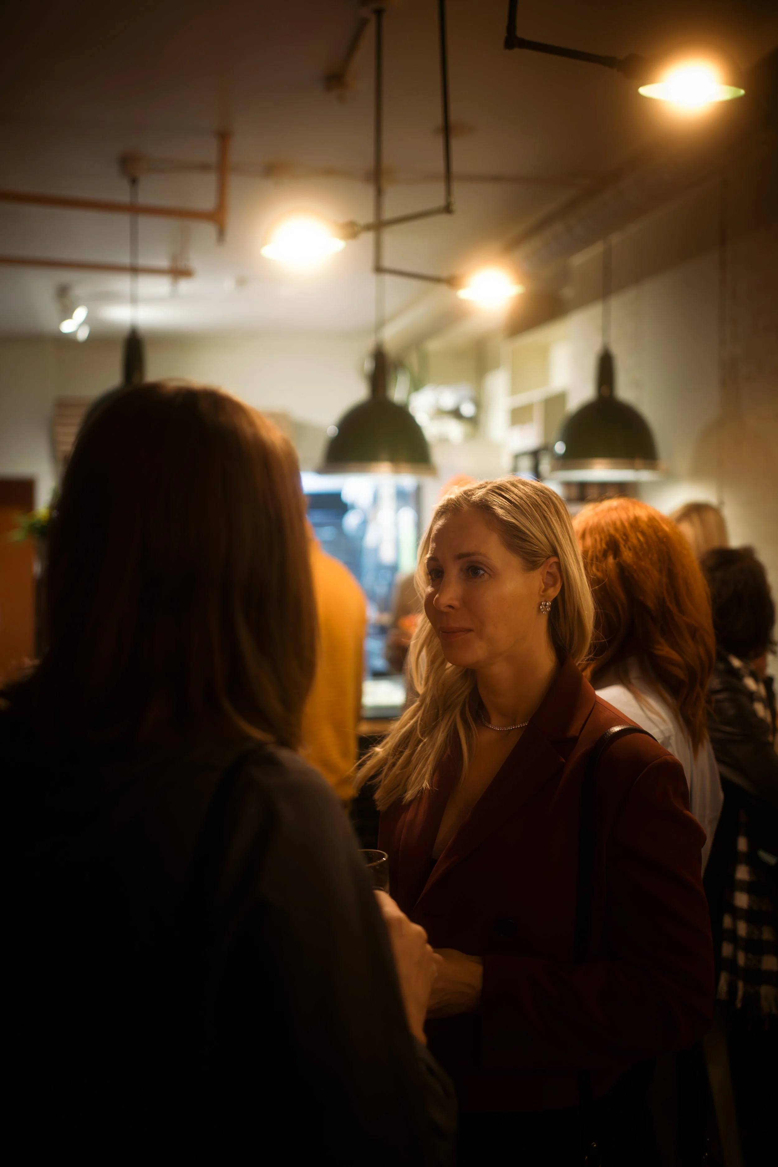 Two women having a conversation in a dimly lit indoor setting, possibly a bar or restaurant, with other people in the background.