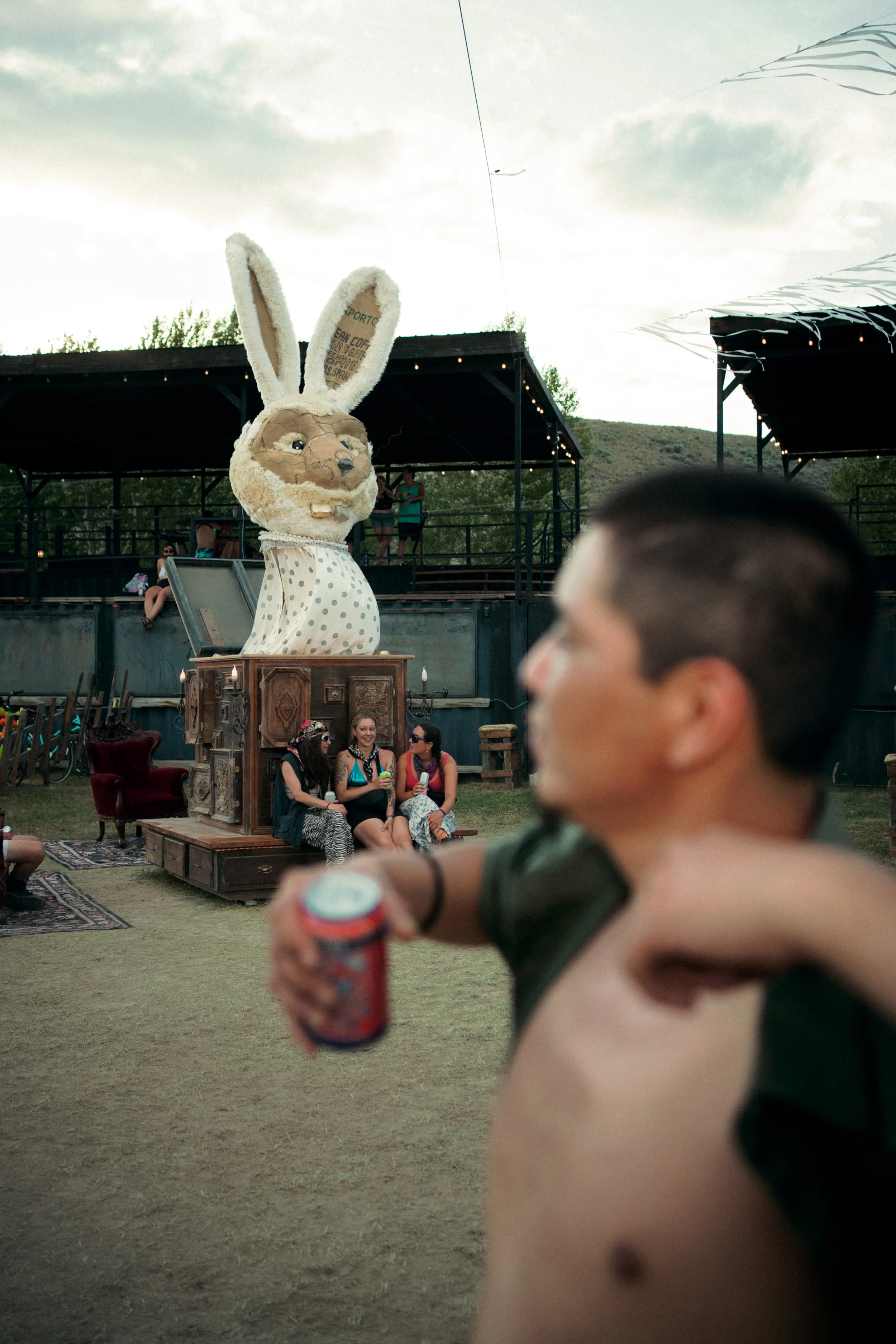 A person holds a soda in the foreground, while in the background there is a large costume of a rabbit with oversized ears, sitting on a vintage cabinet outdoors during the evening, with people sitting on chairs and a stage with a black railing and so