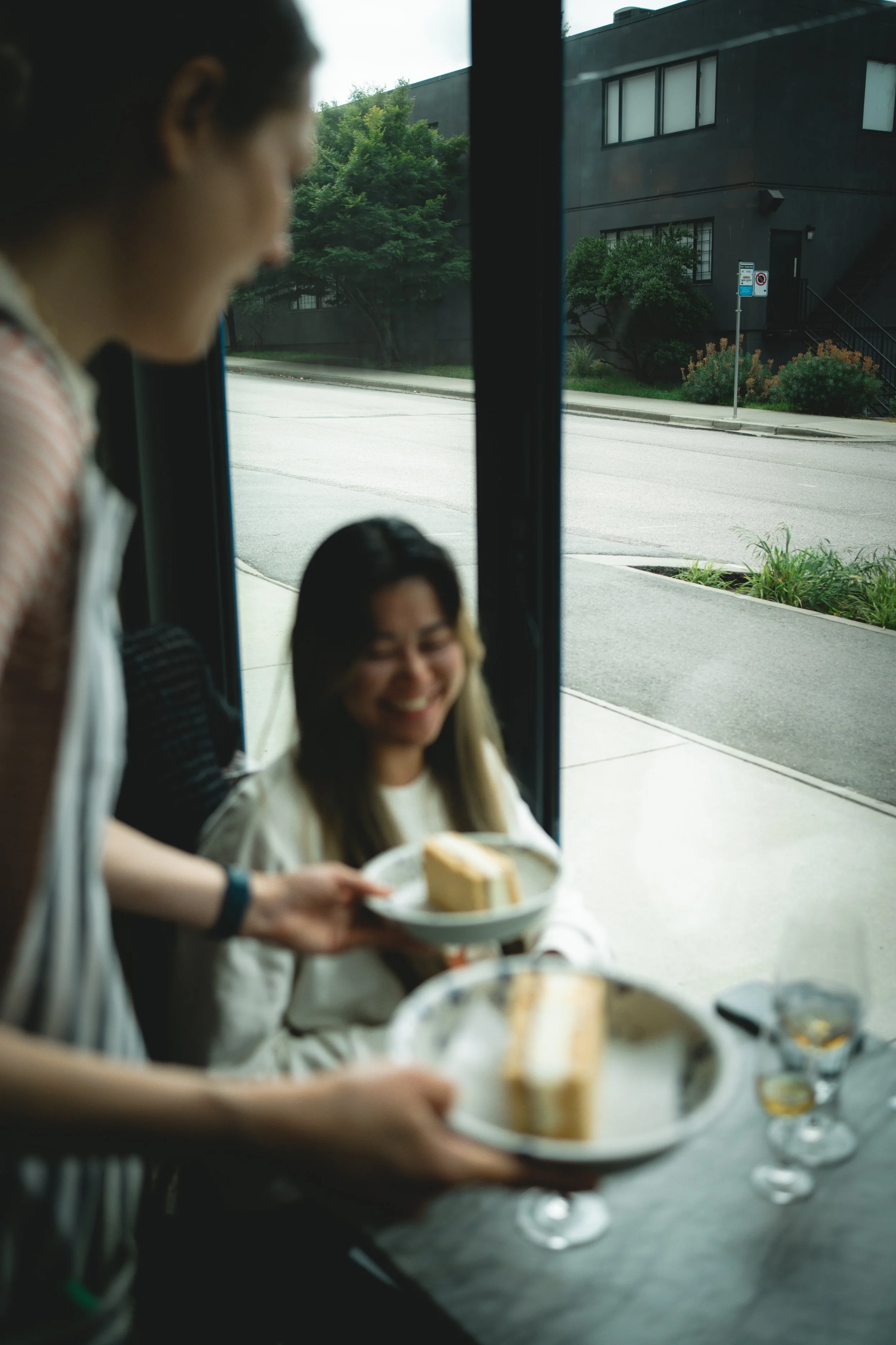 A person inside a cafe serving cake to a woman sitting by the window, who is smiling.