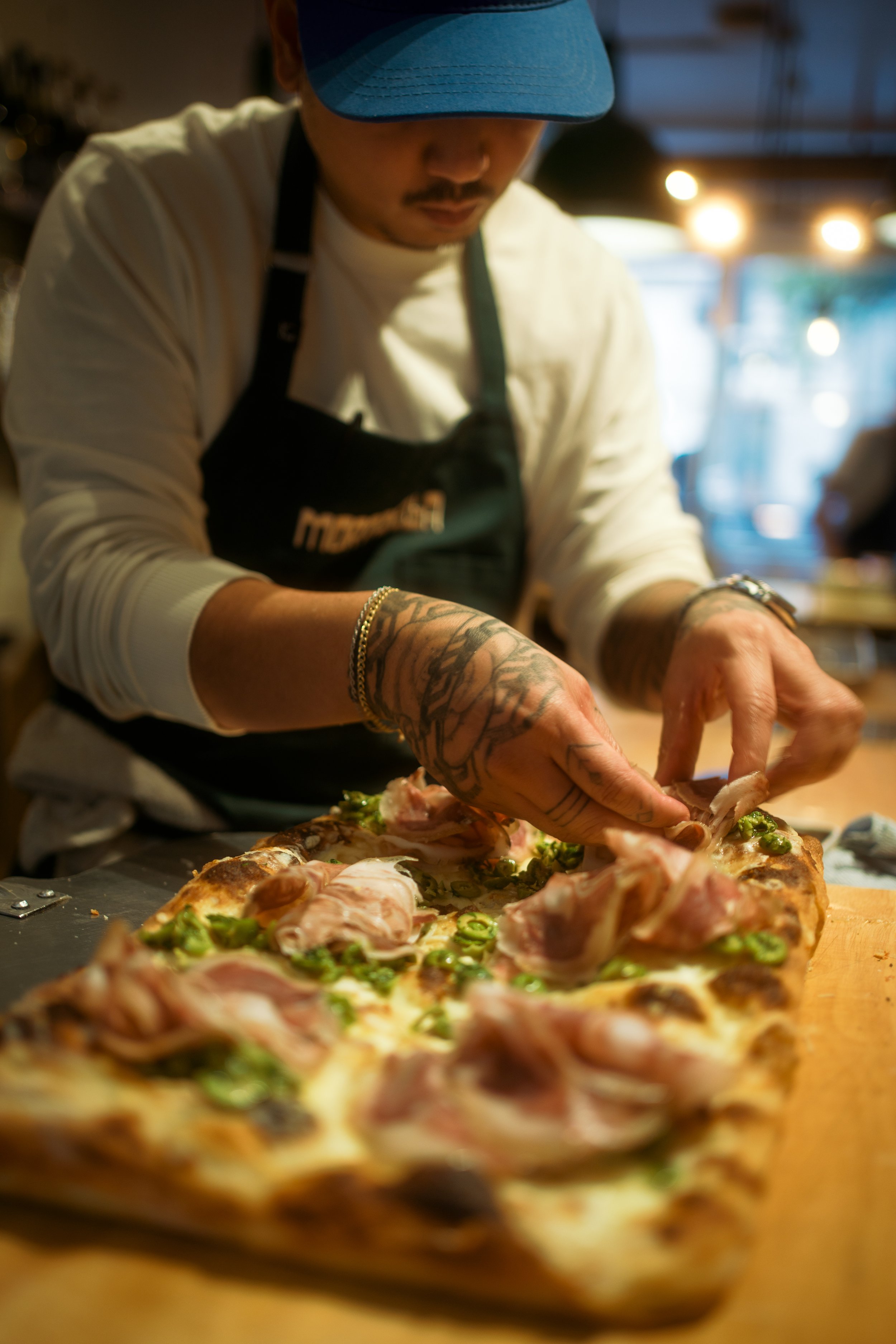 A person with tattoos on their arms, wearing a blue cap, preparing a pizza with chicken and green peppers in a restaurant kitchen.