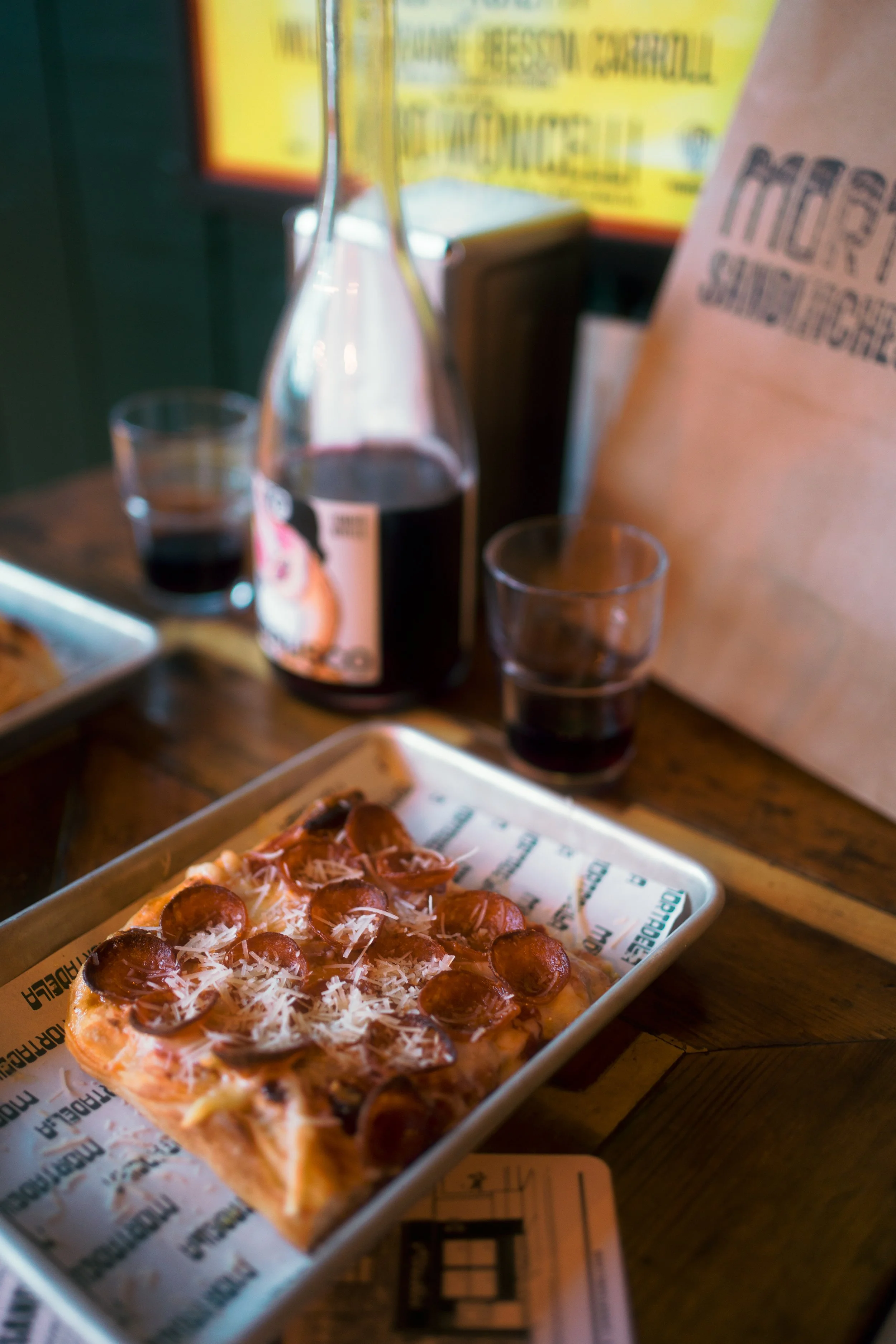 A slice of pepperoni pizza with cheese on a metal tray, two glasses of red wine, a bottle of red wine, a paper bag, a menu, and a plate of bread on a wooden table in a restaurant.