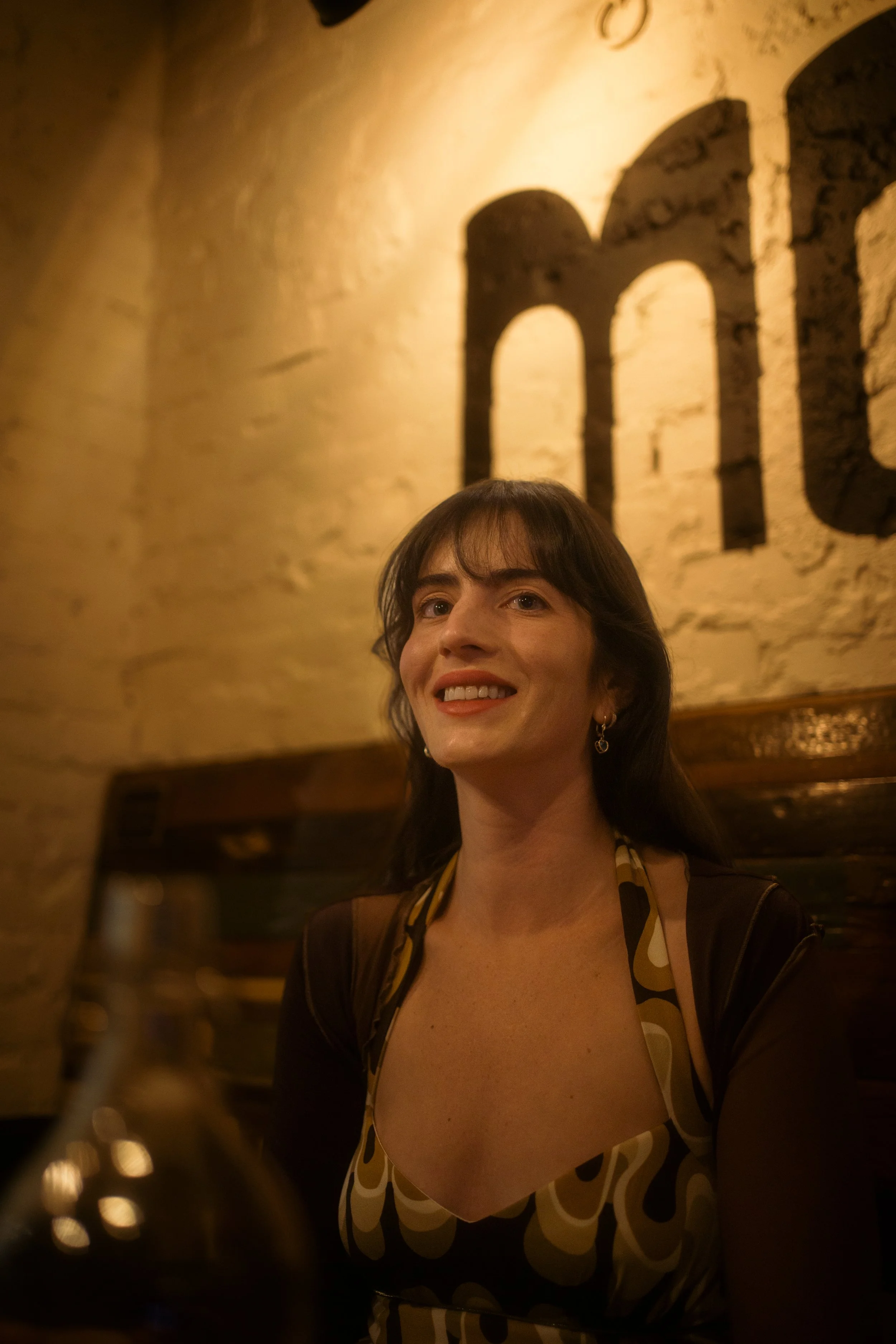 A woman with dark hair and bangs smiling while sitting in a warmly lit restaurant with a stone wall background.